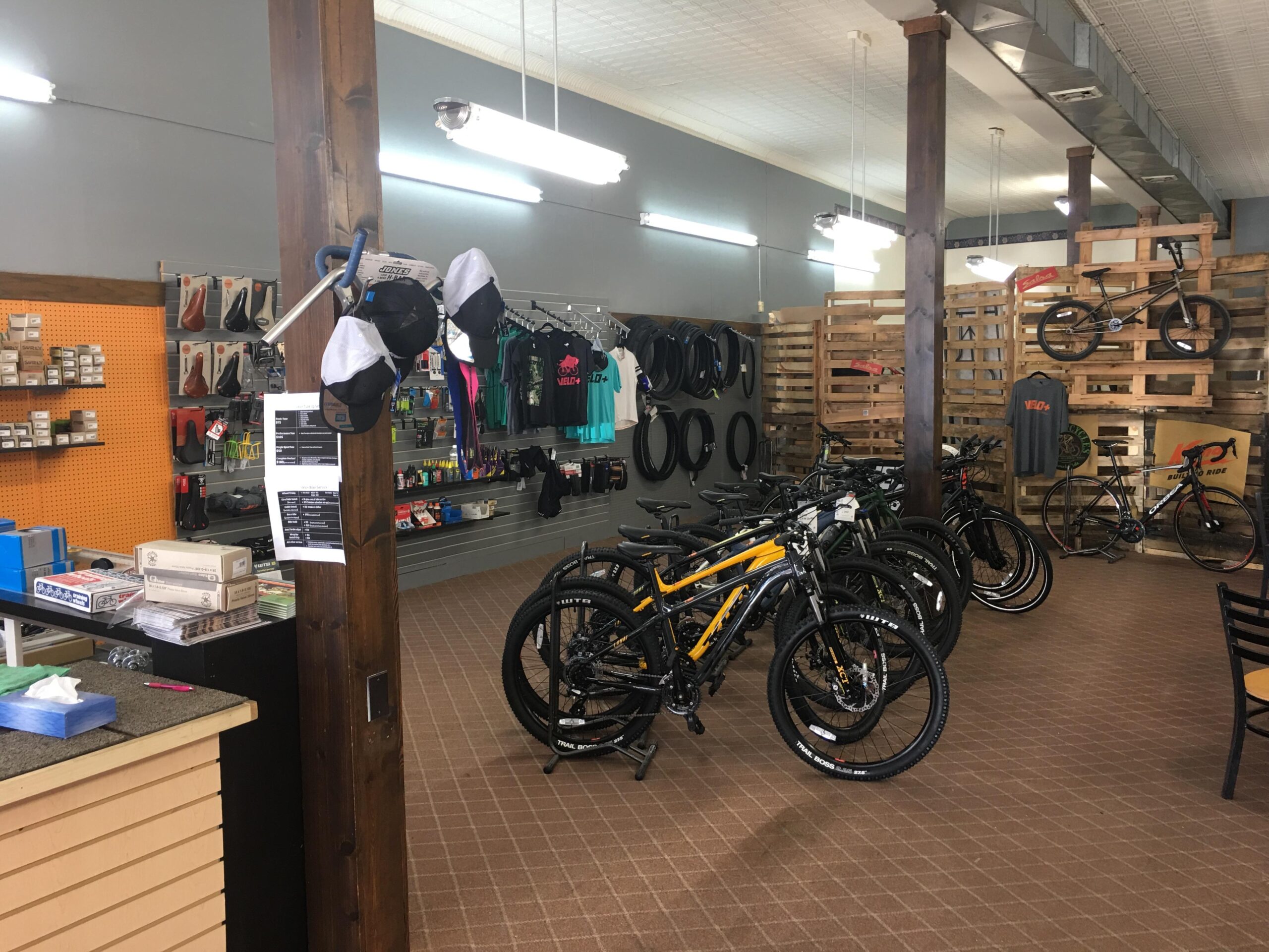 Interior of a bicycle shop featuring several mountain and road bikes lined up on the floor. The shop has wooden beams, display racks with cycling accessories on the walls, and a checkout counter in the foreground. Various cycling apparel items, tires, and tools are visible throughout the store.