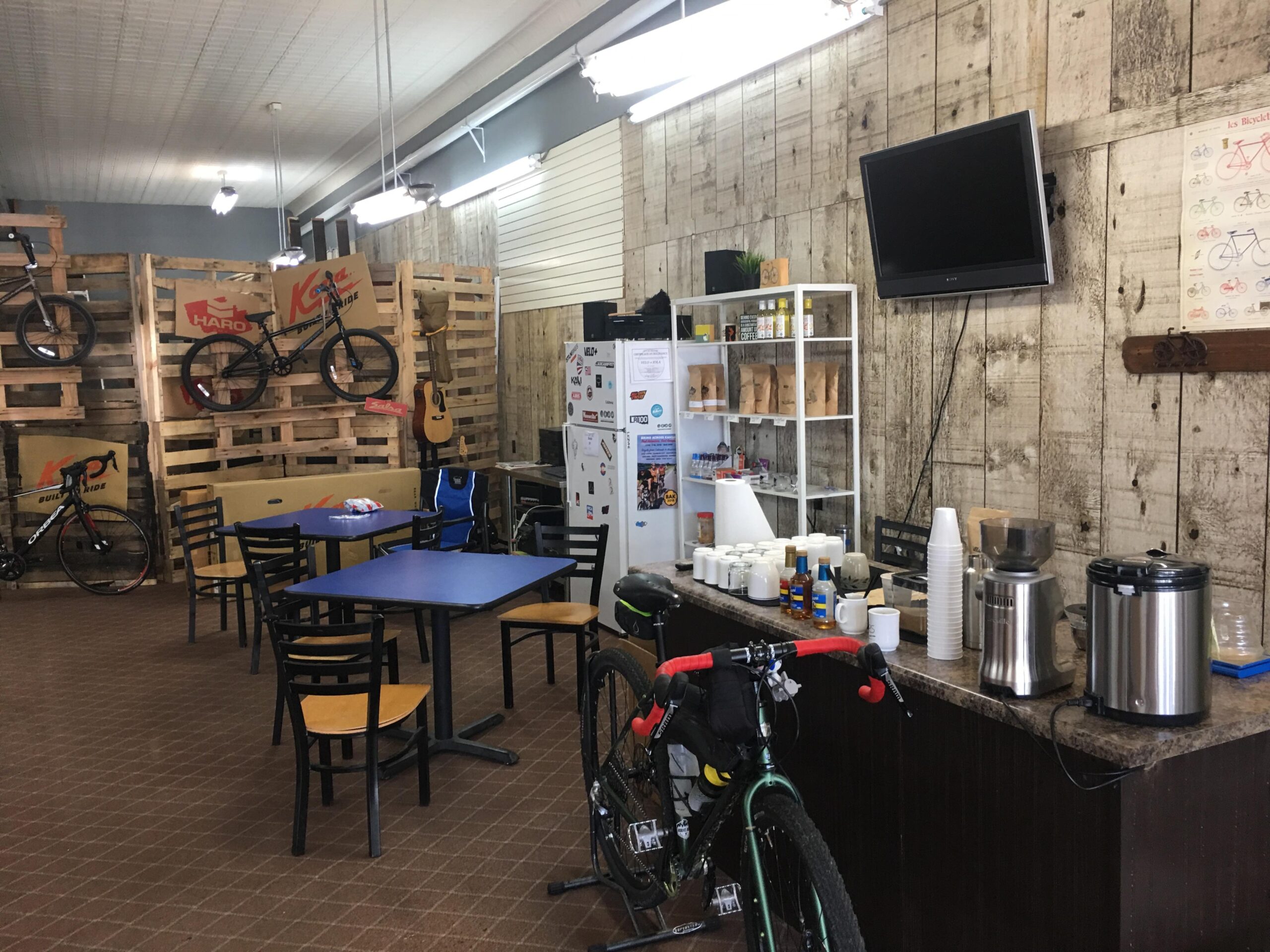 Interior of a cozy bike shop cafe featuring wooden pallets displaying bicycles, a blue table with chairs, a counter with coffee equipment, and a refrigerator. The walls are decorated with wood paneling and a television is mounted on the wall. Various condiments, and snacks are visible on shelves.