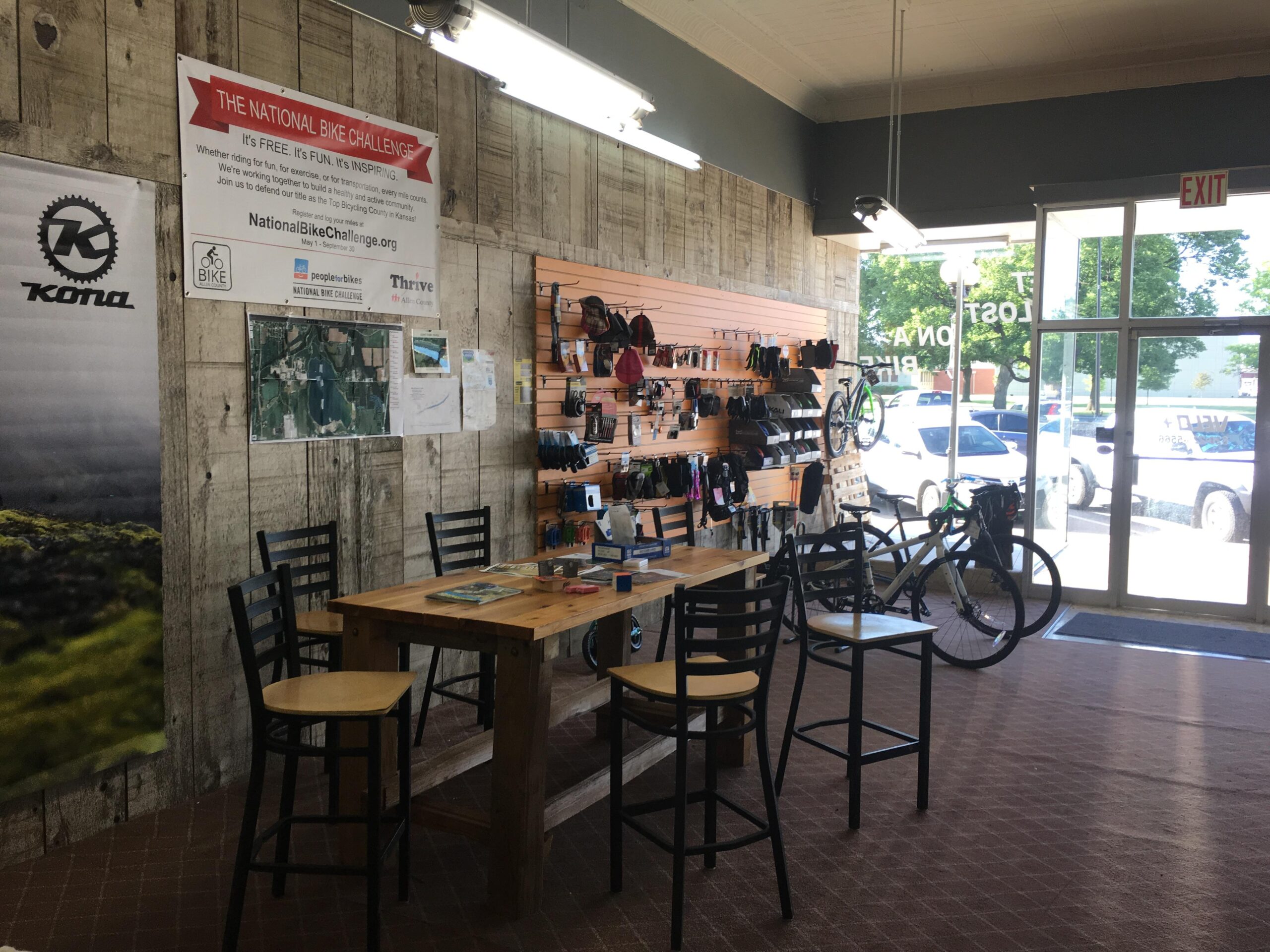 Interior of a bike shop featuring wooden walls, a display with various bike accessories, and a table with chairs. A "National Bike Challenge" banner hangs above. A bike is parked near the entrance, with natural light coming in from the window.