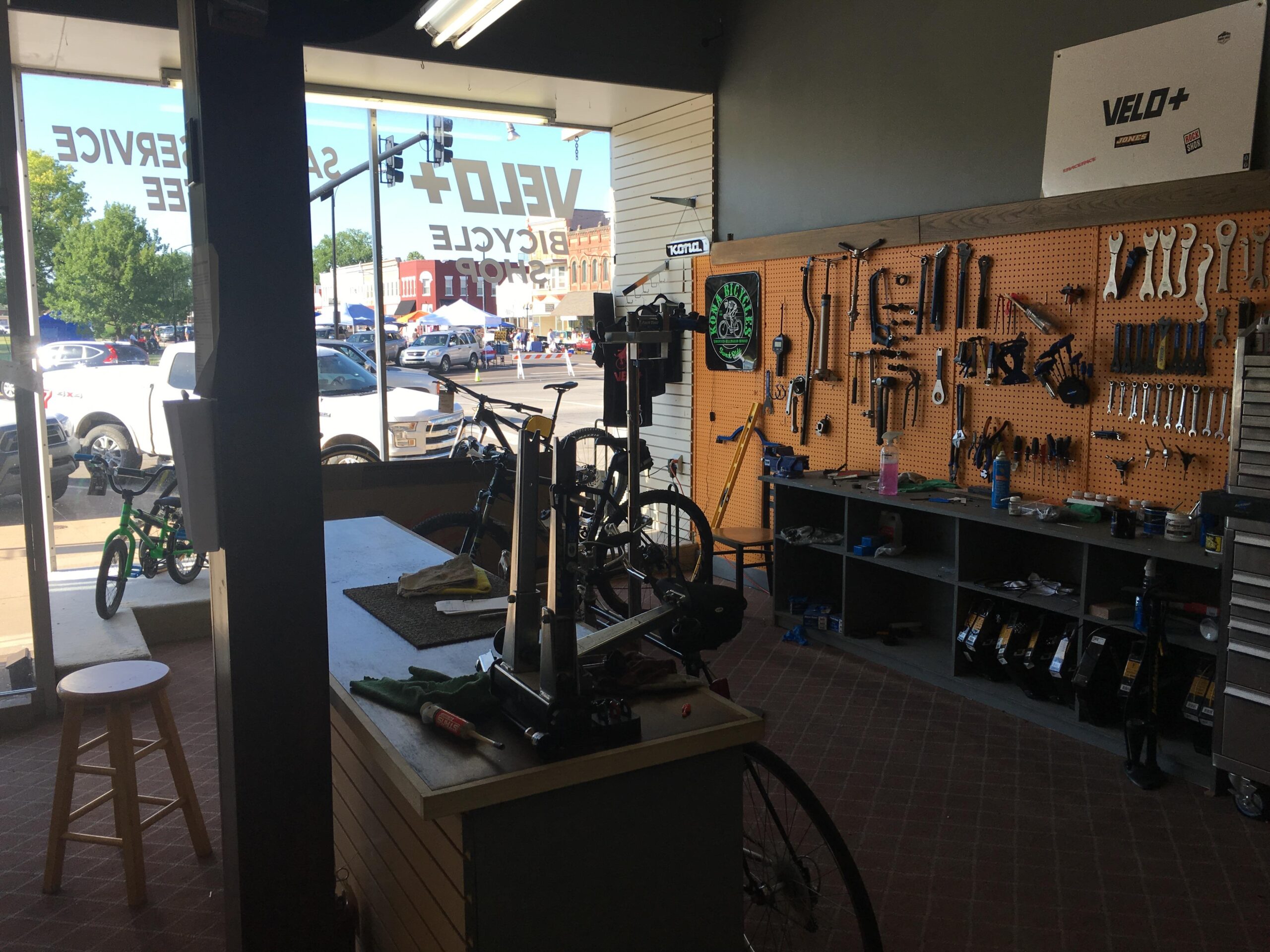 Interior of a bicycle shop featuring a workbench with tools and bike repair equipment. The wall behind has organized tools hanging on a pegboard. A view of the street through large windows shows parked vehicles and people outside. A stool is placed near the workbench.