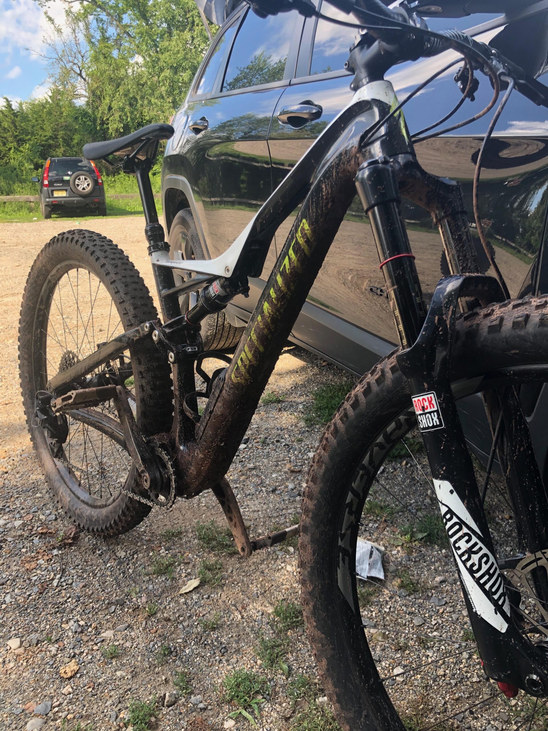 Specialized Stumpjumper FSR 6Fattie: A close-up view of a muddy mountain bike leaning against a parked black SUV. The bike features robust tires and a prominent suspension system, indicating recent use on rugged terrain. In the background, there are green trees and a gravelly surface.