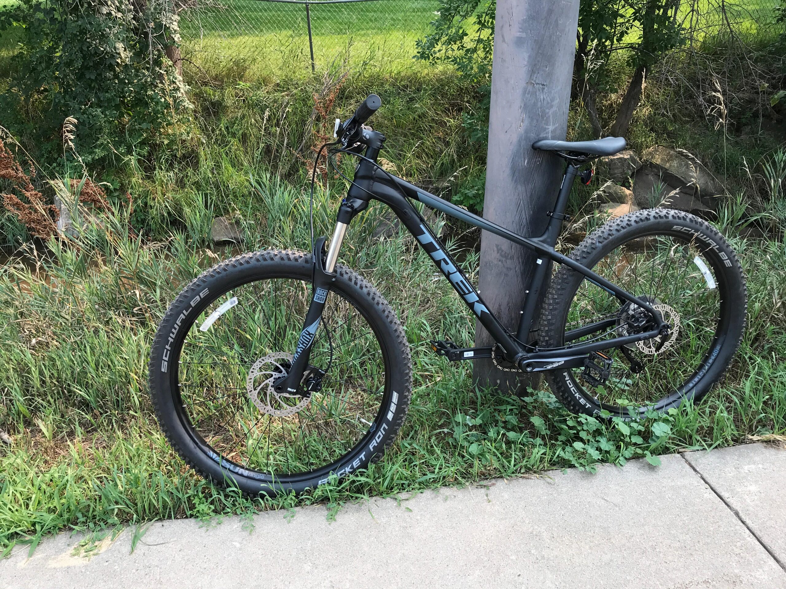 Trek Roscoe 7: A black Trek mountain bike leaning against a wooden post, surrounded by green grass and foliage. The bike features thick tires designed for off-road use, with visible branding on the tires and frame. In the background, a fence and a grassy area are visible, indicating a natural setting.