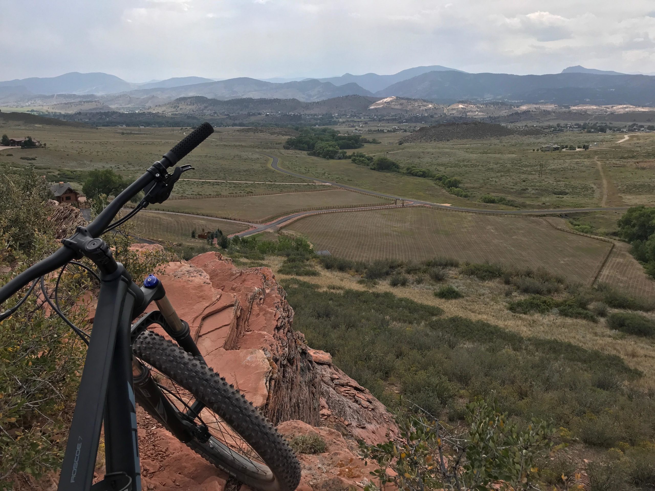 A mountain bike rests on a rocky outcrop, overlooking a vast, green landscape with rolling hills and distant mountains under a cloudy sky. The foreground shows the bike's handlebar and tire, while the background features farmland and winding roads. Blue Sky mountain bike trail.