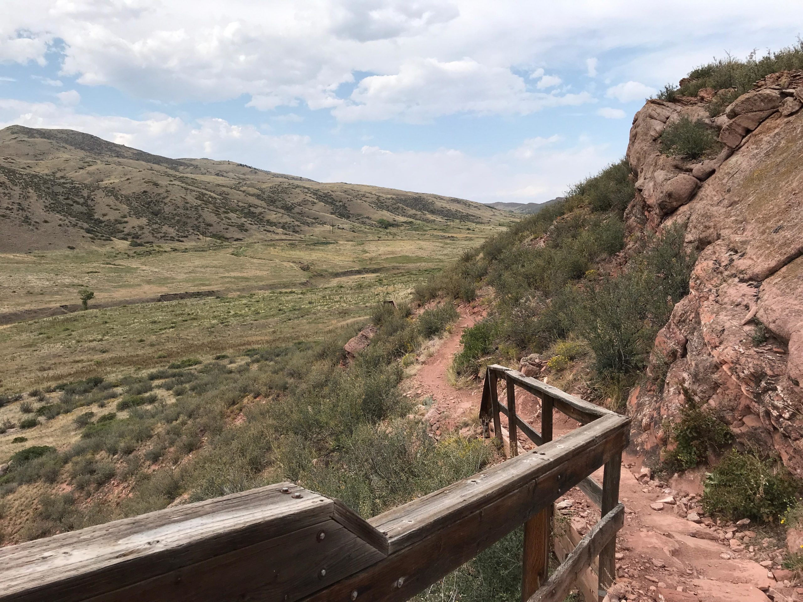 A winding dirt path along a hillside with a wooden railing, surrounded by sparse vegetation and rocky terrain. In the background, rolling hills stretch under a partly cloudy sky, showcasing a serene natural landscape. Blue Sky mountain bike trail.