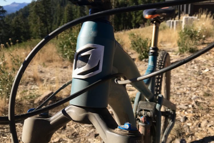 Close-up of a mountain bike's frame showcasing the head tube with a logo, cable routing, and front suspension fork. The background features a natural setting with trees and dry grass, suggesting an outdoor environment.