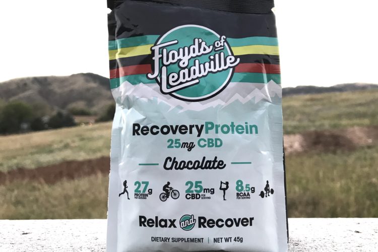 A bag of Floyd's of Leadville Recovery Protein in chocolate flavor, featuring 25 mg of CBD, with details highlighting 27 g of protein and 8.5 g of BCAA per serving. The background shows a scenic outdoor landscape with rolling hills.