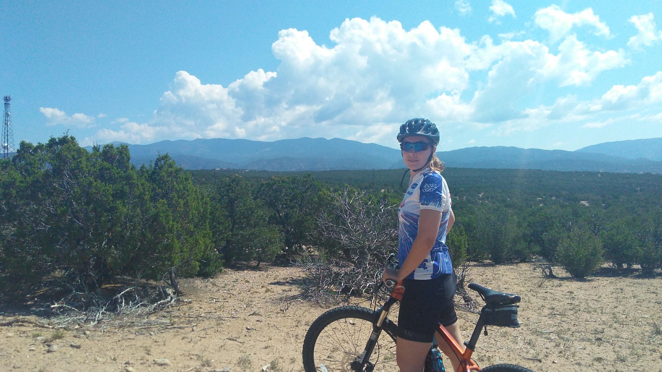 A mountain biker wearing a blue and white jersey, sunglasses, and a helmet, standing next to an orange mountain bike in a rocky landscape with bushes and mountains in the background. The sky is blue with fluffy clouds. La Tierra mountain bike trail.