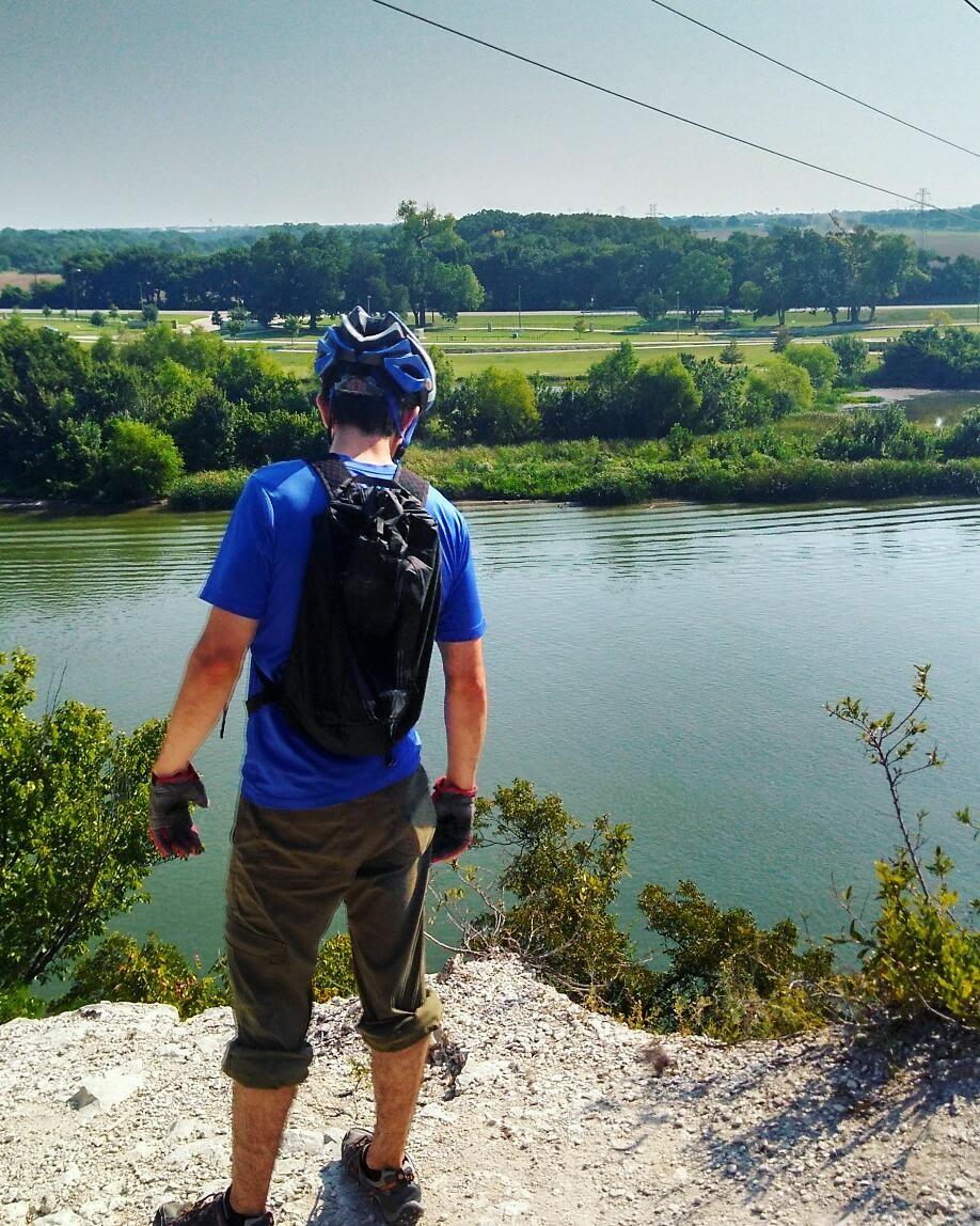 A person wearing a blue helmet and gloves stands at the edge of a rocky outcrop, looking out over a calm river surrounded by greenery. The scene is set on a sunny day, with trees and a park visible in the background. Cameron Park mountain bike trail.