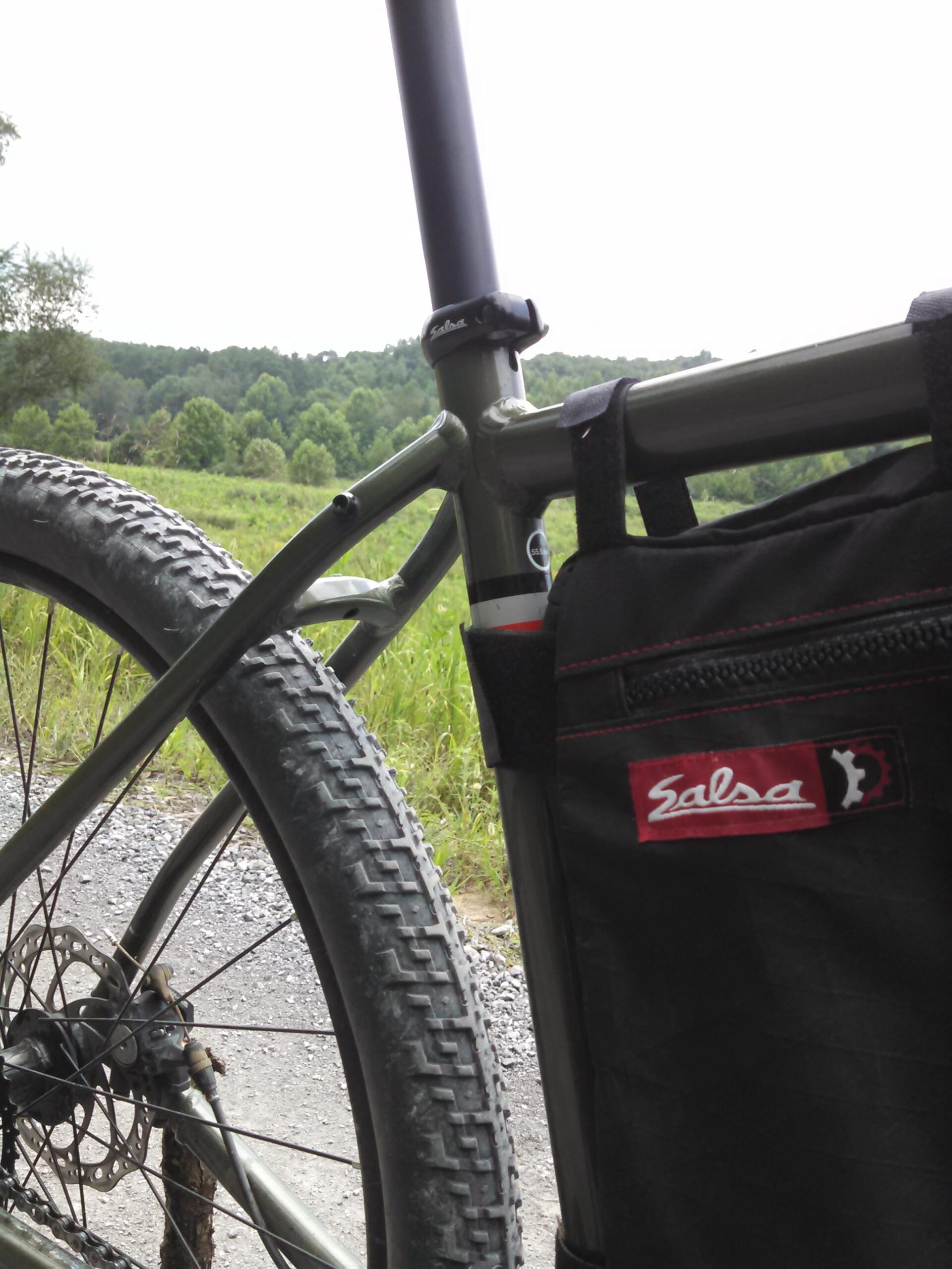 Salsa Jouneryman: A close-up view of a bicycle frame and rear wheel, featuring a Salsa bag attached to the frame. The background includes grassy fields and a forested hillside under a cloudy sky.