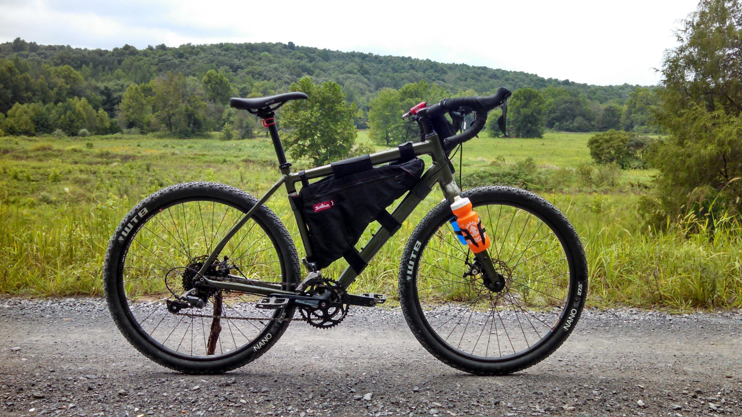 Salsa Jouneryman: A side view of a bike positioned on a dirt path, set against a backdrop of a green, hilly landscape under a cloudy sky. The bike features a black frame, wide tires, and a black bike bag attached to the frame. An orange water bottle is secured in a holder on the frame.