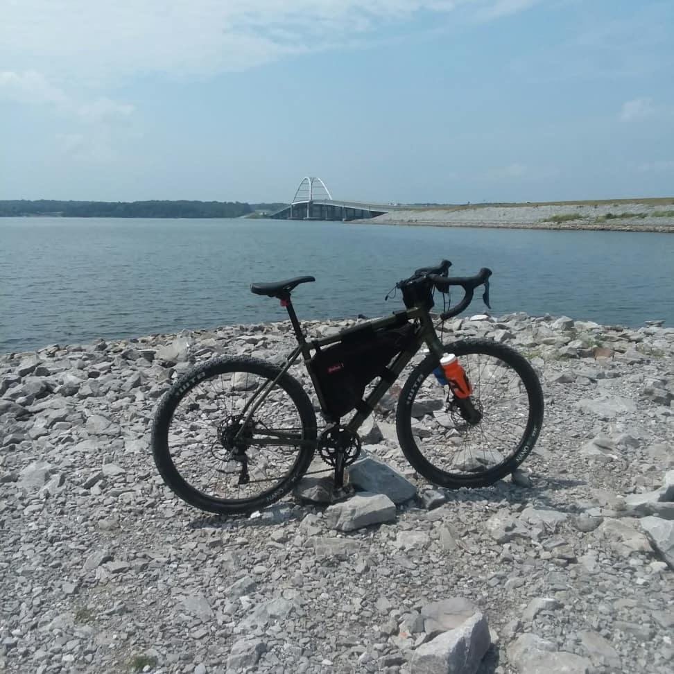 Salsa Jouneryman: A black bicycle is parked on rocky terrain by a body of water, with a bridge visible in the background. The scene is set under a clear blue sky, suggesting a sunny day outdoors.