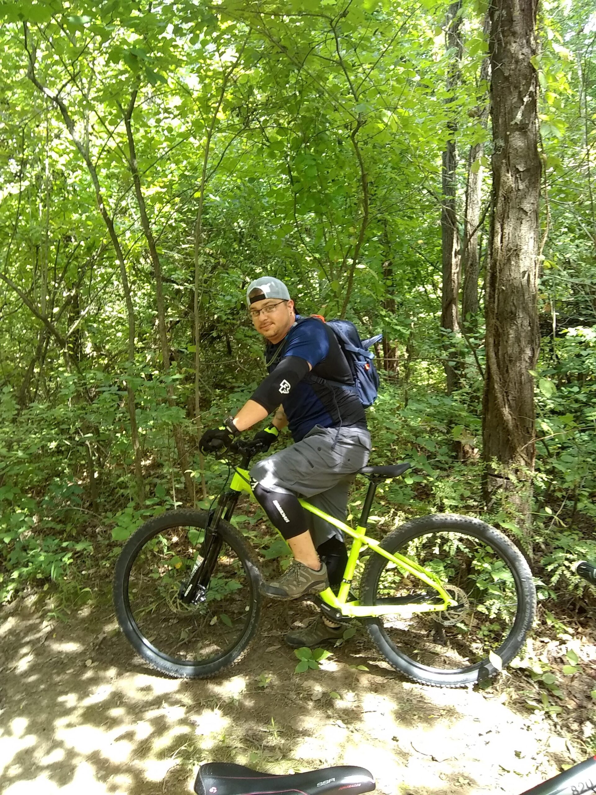 Trek Marlin 5: A cyclist resting on a bright yellow mountain bike in a lush green forest. The rider is wearing a blue shirt, gray shorts, and safety gloves, with a backpack and a cap. Surrounding them are dense trees and foliage, creating a natural, outdoor setting.