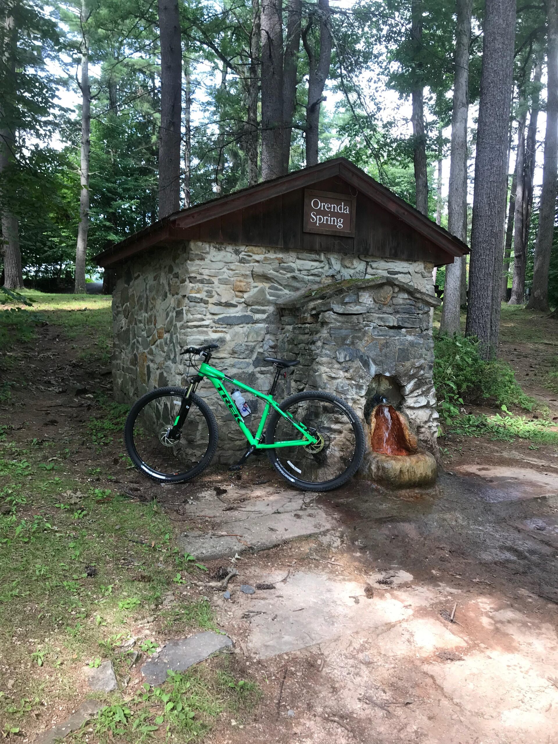 Trek Marlin 5: A green mountain bike rests against a stone structure labeled "Orenda Spring," with a small water spring flowing nearby. Surrounded by lush trees, the scene depicts a peaceful natural setting.