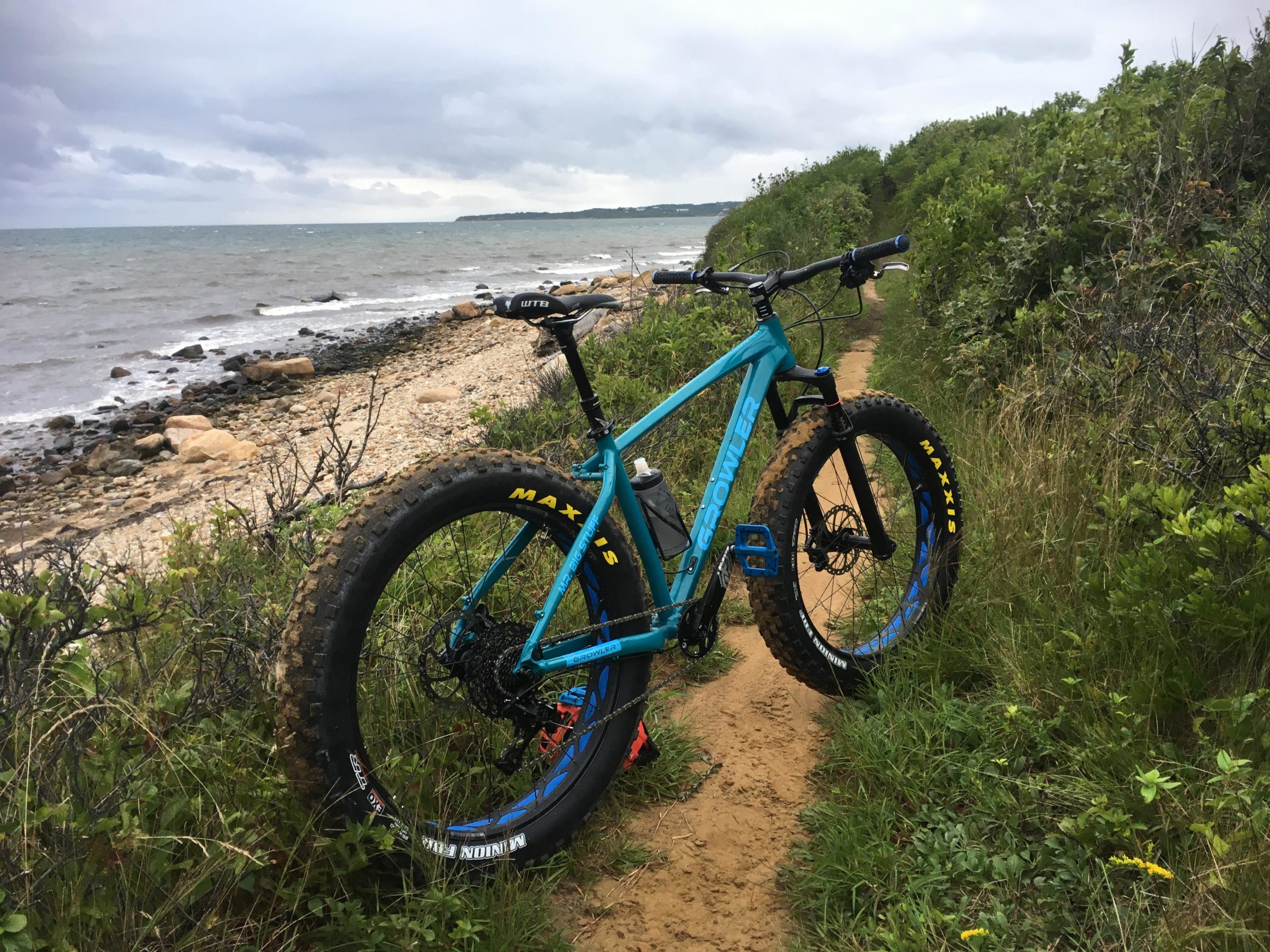 A bright blue fat bike is parked along a sandy trail near the shoreline. The landscape features a rocky beach with waves lapping at the shore, surrounded by lush green vegetation. Overcast skies are visible above, suggesting a cool day. Hither Woods mountain bike trail.