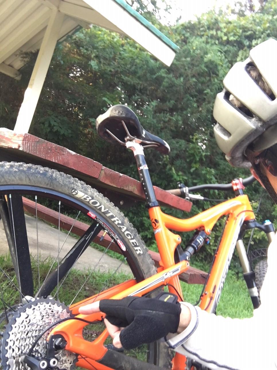 Santa Cruz Tallboy Carbon: A close-up view of a cyclist's hand adjusting the drivetrain of an orange mountain bike, featuring a Bontrager tire. The scene is set near a wooden structure with green foliage in the background. The cyclist is wearing a helmet and gloves.