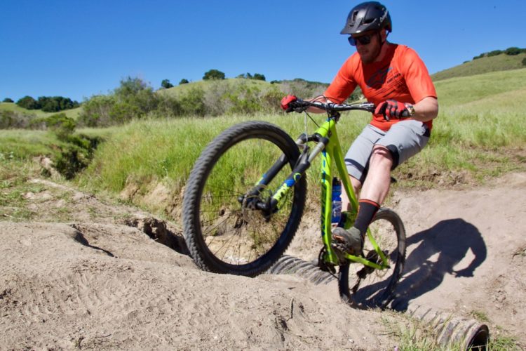 A mountain biker in an orange shirt and helmet performs a jump on a dirt trail, with one wheel elevated above a rocky section. Lush green hills and a clear blue sky serve as the backdrop, showcasing a sunny day outdoors.