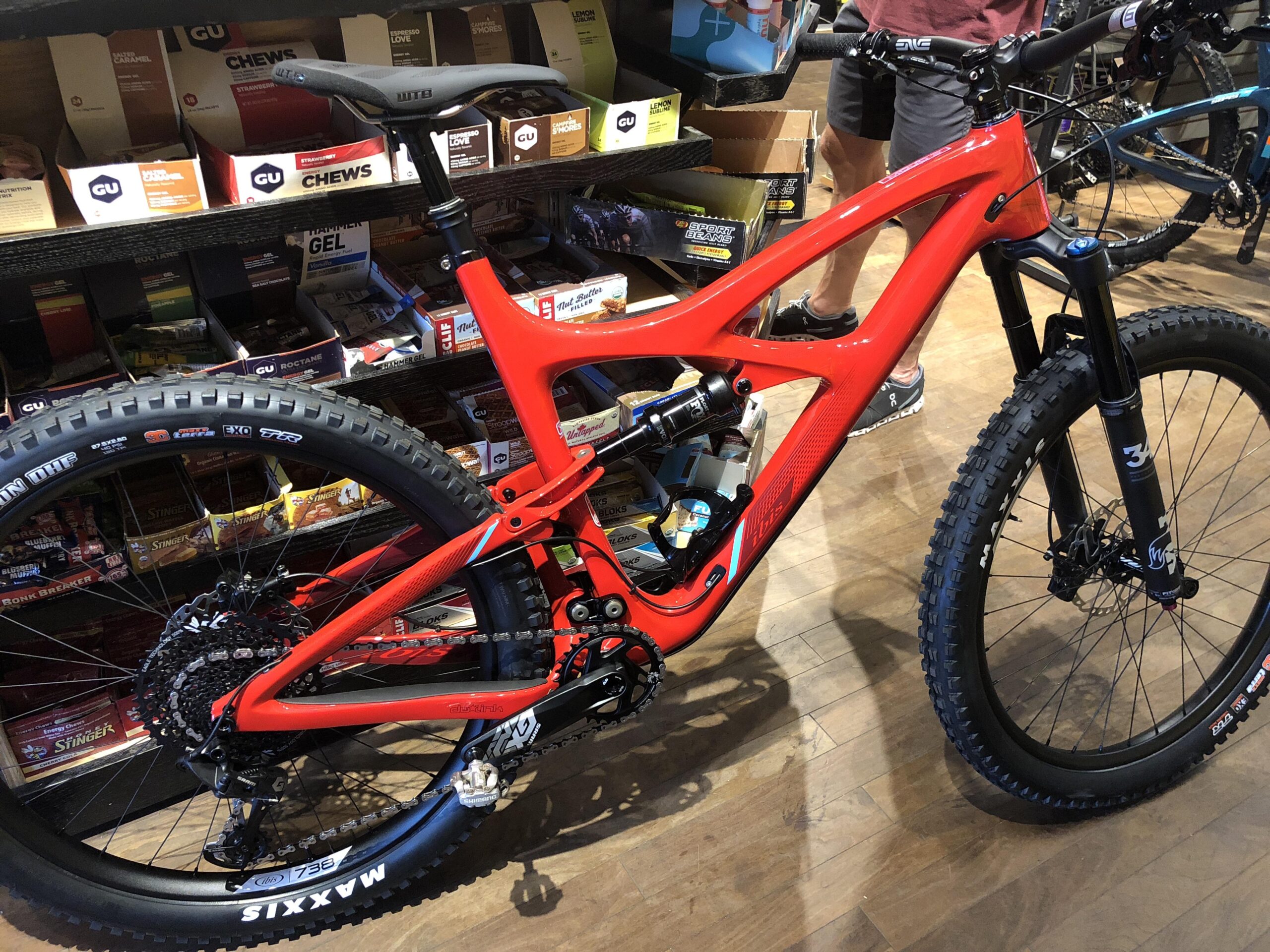 Ibis Mojo 3: A bright red mountain bike is displayed in a store, showcasing its sleek frame and design. The bike features thick tires labeled "Maxxis" and detailed components, including a visible drivetrain and front suspension. In the background, various energy bars and gels are neatly arranged on shelves, indicating a focus on outdoor sports and biking accessories.