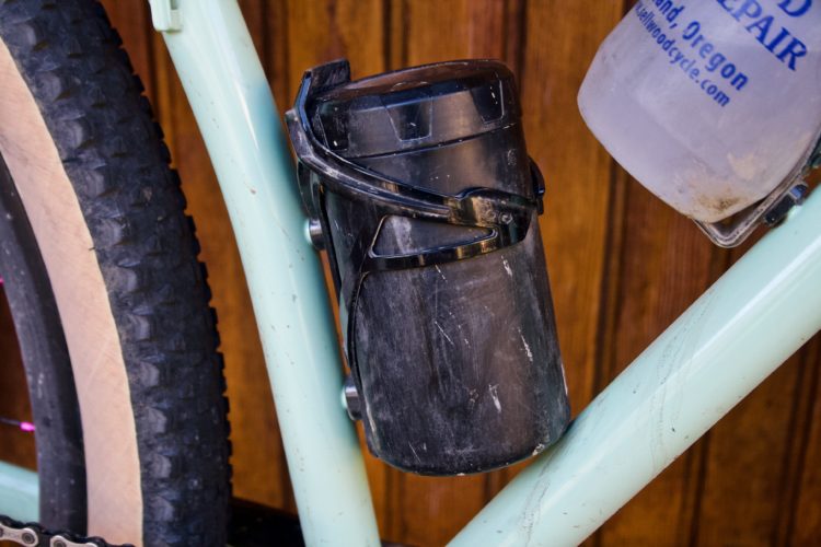 Close-up of a mountain bike frame featuring a black water bottle holder attached to the side, with a partially visible clear water bottle above it. The bike has textured tires and a mint green frame, set against a wooden background.