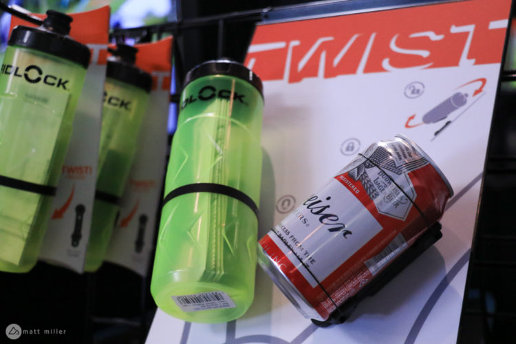 A display featuring a green bicycle water bottle labeled "BLOCK" and a can of beer on a promotional board with the word "TWIST." The background includes diagrams illustrating the use of the product.