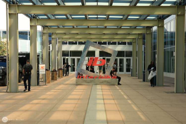 An outdoor view of the Interbike event entrance, featuring a large metal sculpture with the logo "iB" and the word "Interbike" prominently displayed. People are seen walking along the walkway beneath a glass-covered structure, and there are event booths and displays in the vicinity.