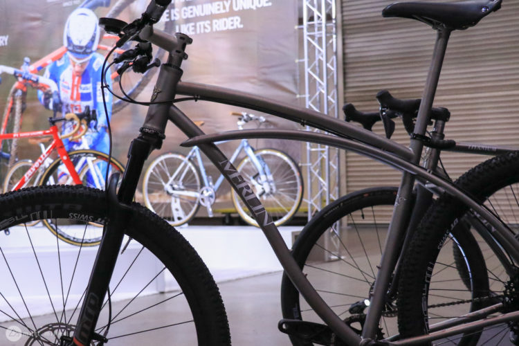 A close-up view of a mountain bike in a display setting, showcasing its sleek frame and sturdy tires. In the background, other bicycles can be seen, along with a promotional banner featuring a cyclist. The overall atmosphere suggests a focus on cycling gear and design.