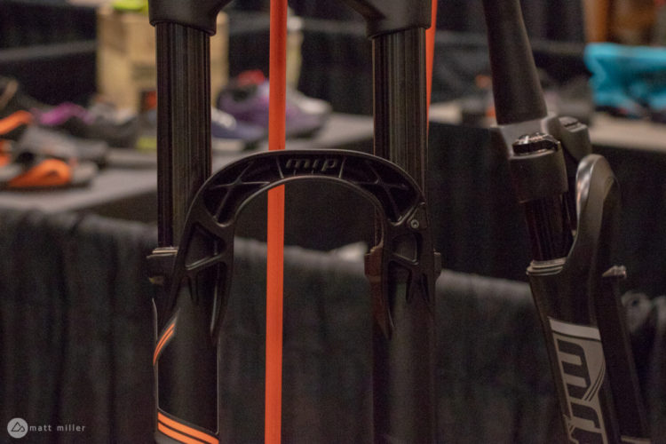 Close-up of a mountain bike fork, featuring a black and orange design. The fork's bridge and legs are visible, with a blurred background showing other biking accessories.