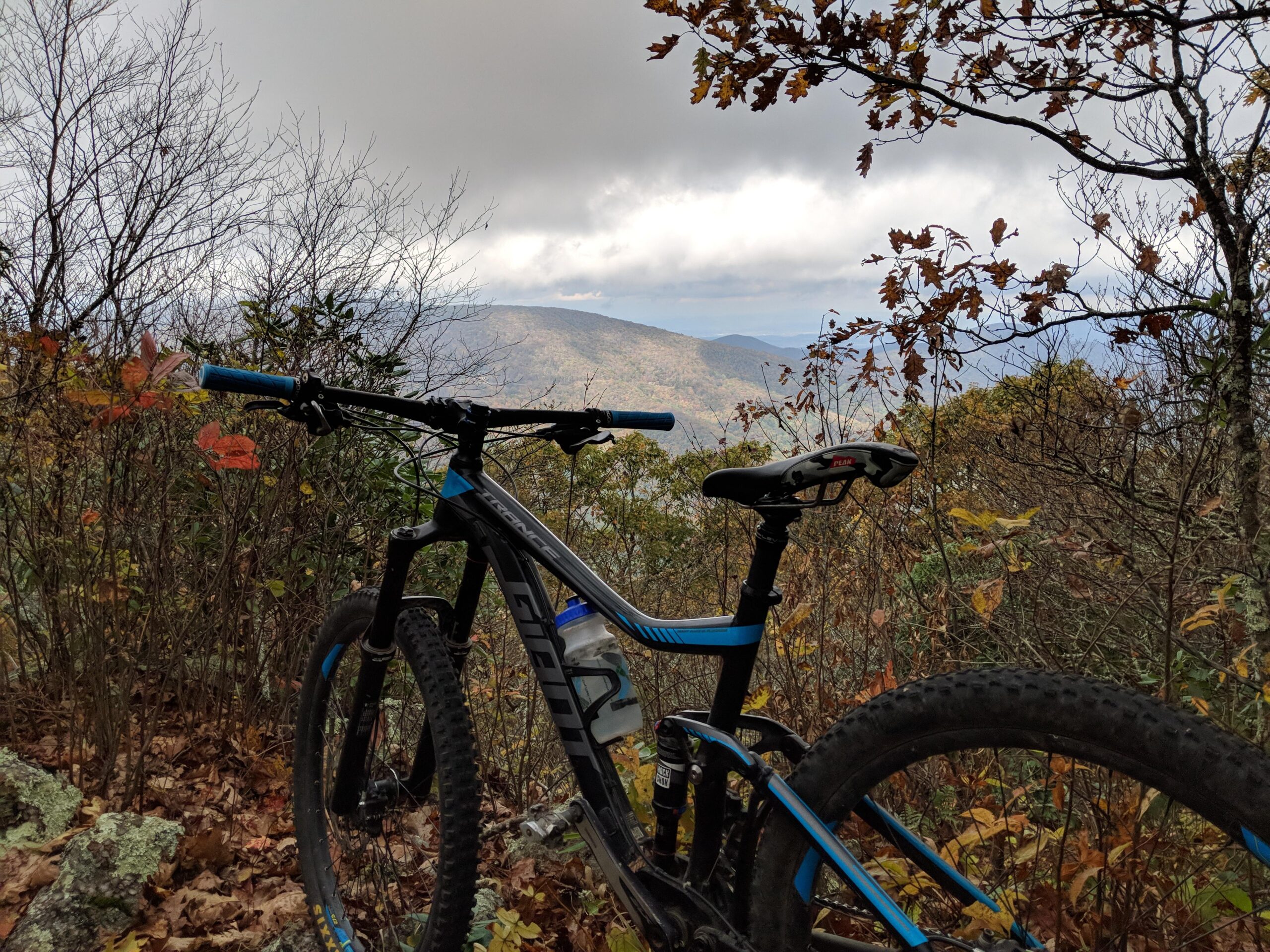 Giant Trance: A mountain bike is positioned on rocky terrain, surrounded by trees and autumn foliage, with distant mountains and cloudy skies visible in the background.