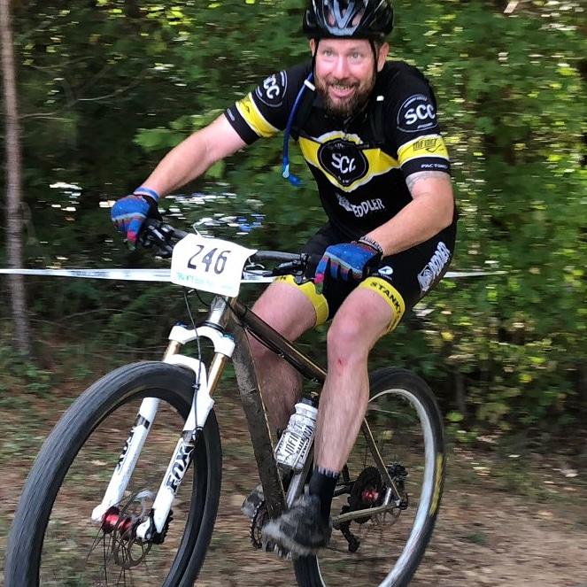 A smiling cyclist wearing a black and yellow jersey and shorts rides a mountain bike on a wooded trail. The bike has a visible race number (246) attached to the front. The scene depicts a lively, outdoor activity with greenery in the background. Montgomery Bell State Park Mtb Trail mountain bike trail.