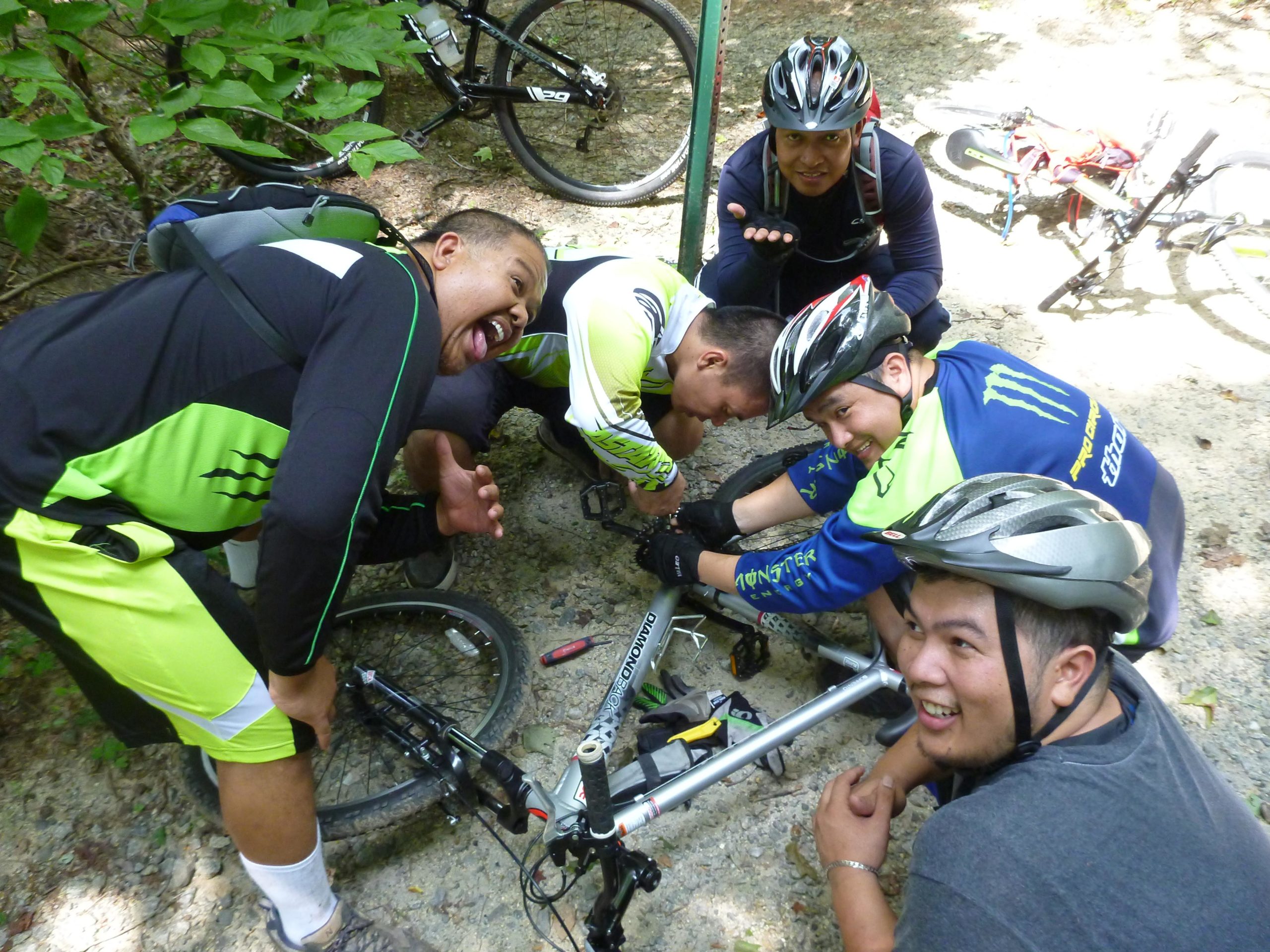 A group of five men wearing cycling helmets and bright jerseys gather around a bicycle on a dirt path, laughing and engaging in a playful moment. One man is crouching and making a funny face, while another leans in to examine the bike. Tools are scattered nearby, suggesting they are fixing the bike together amidst a natural, leafy backdrop. Wild Turkey mountain bike trail.