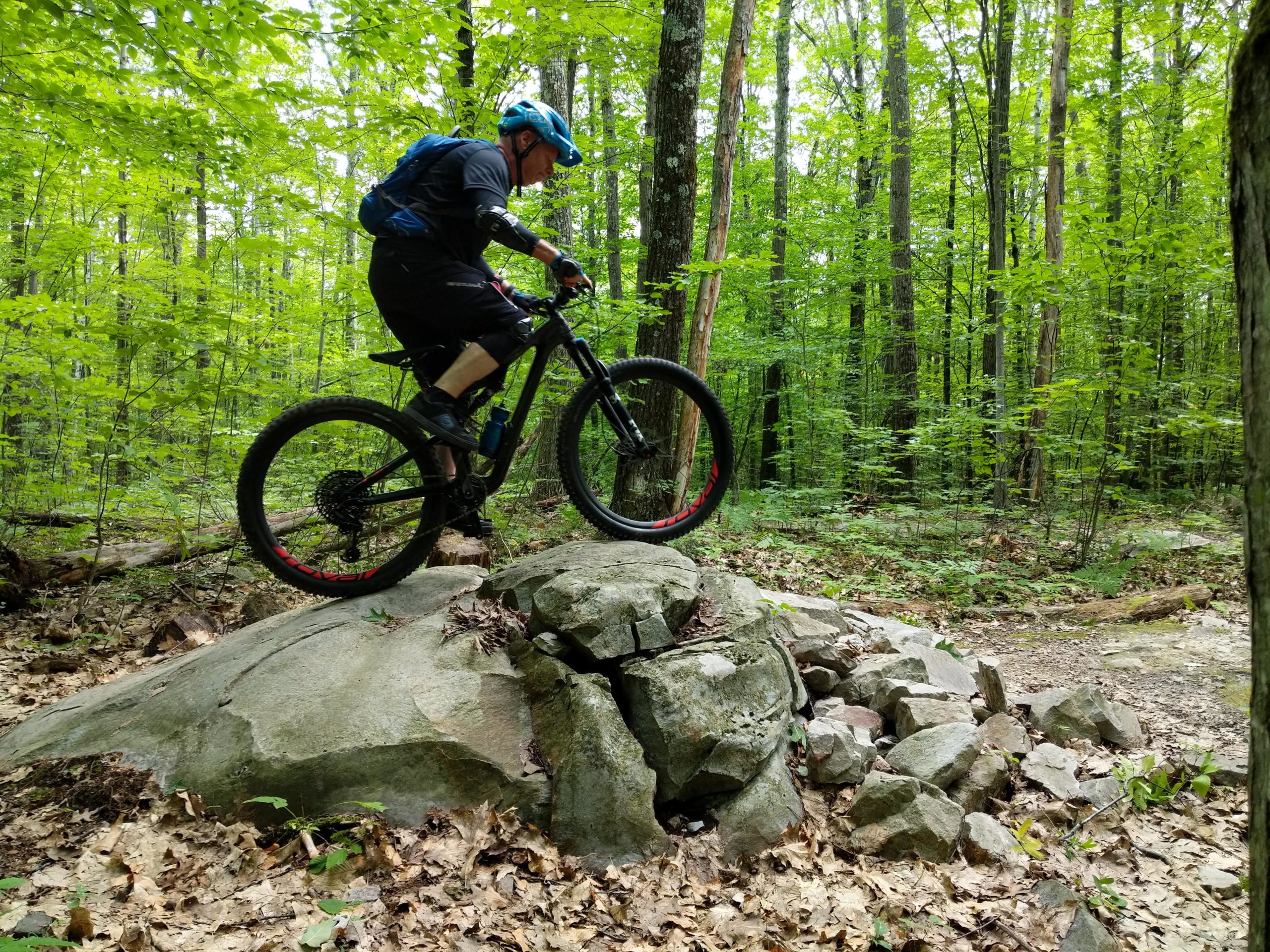 A mountain biker navigating a rocky terrain in a lush, green forest. The cyclist is balancing on a large boulder, showcasing skills in an outdoor setting surrounded by trees and scattered leaves. Pine Hill Park mountain bike trail.