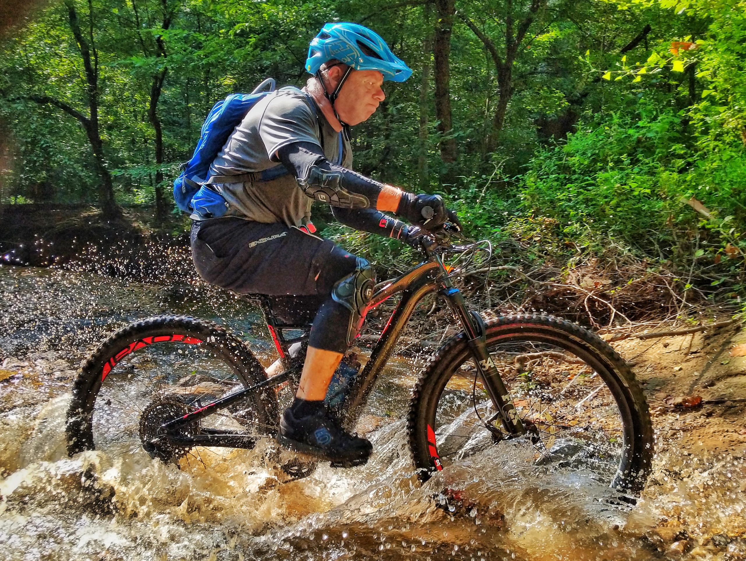 A mountain biker splashes through water on a dirt trail surrounded by lush greenery. The rider, wearing a blue helmet and protective gear, demonstrates skill and excitement as water sprays around the bike's wheels. The scene captures the thrill of outdoor adventure in nature. Patapsco Valley State Park (Avalon Area) mountain bike trail.