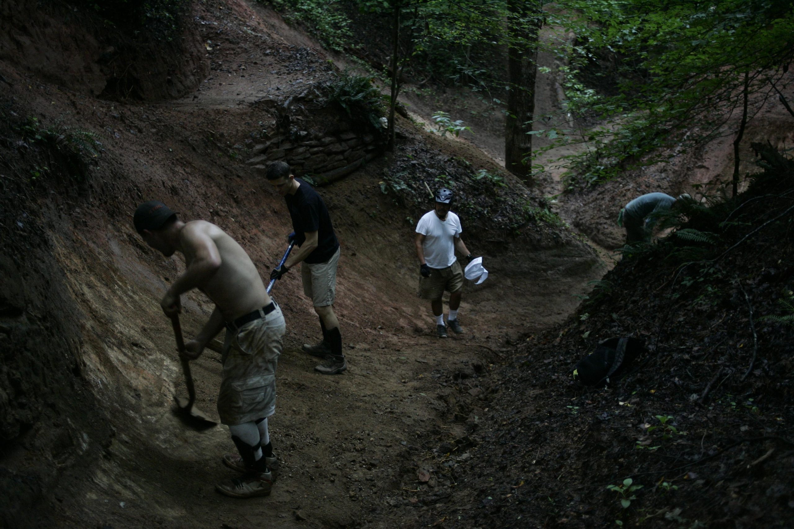 Groups of people working in a wooded area, using shovels and tools to dig and shape the trails. The scene is set in a natural environment with visible earth and vegetation. A mix of individuals is wearing various clothing styles, including some in shorts and t-shirts, as they collaborate on outdoor labor. Salem Lake mountain bike trail.