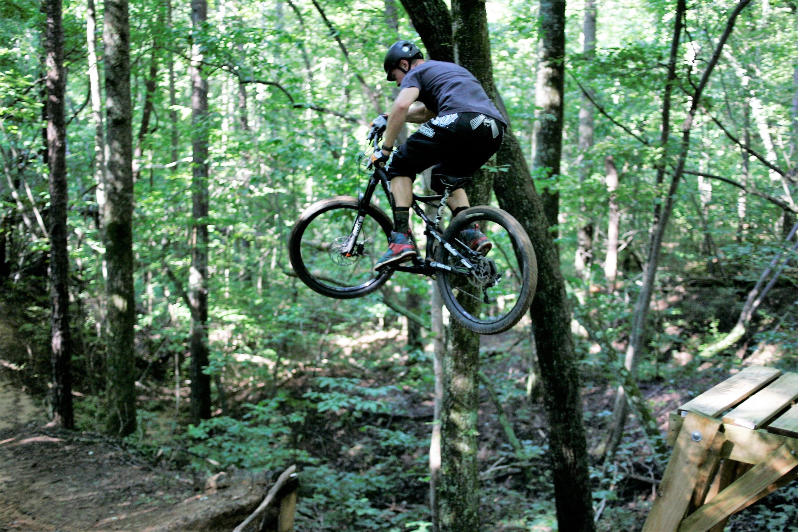 A mountain biker performing a jump over a wooden ramp while riding through a dense forest, surrounded by lush green trees. The biker is airborne, showcasing a dynamic mid-air position. Salem Lake mountain bike trail.