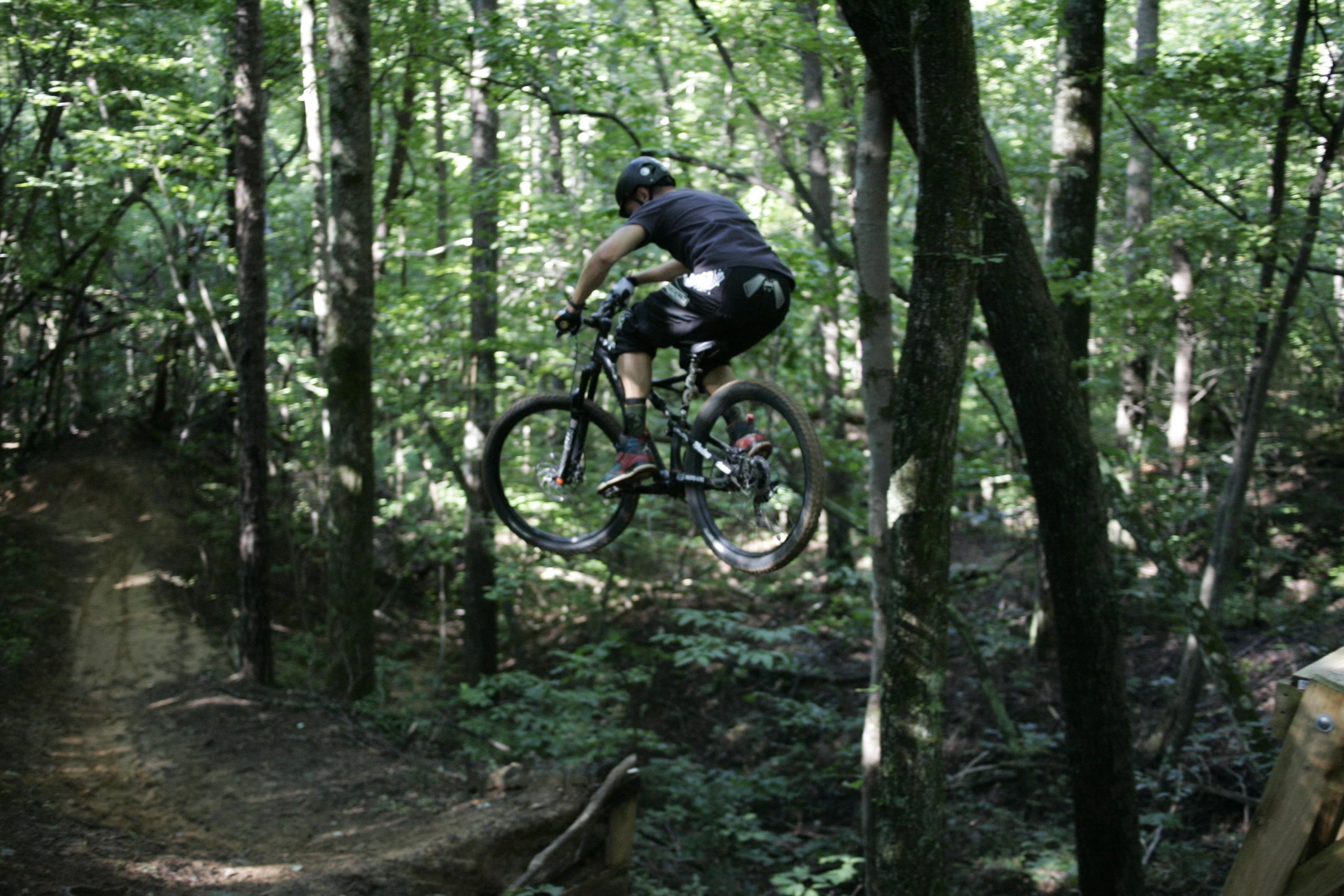 A mountain biker performing a jump over a dirt ramp in a lush green forest, surrounded by trees. Salem Lake mountain bike trail.