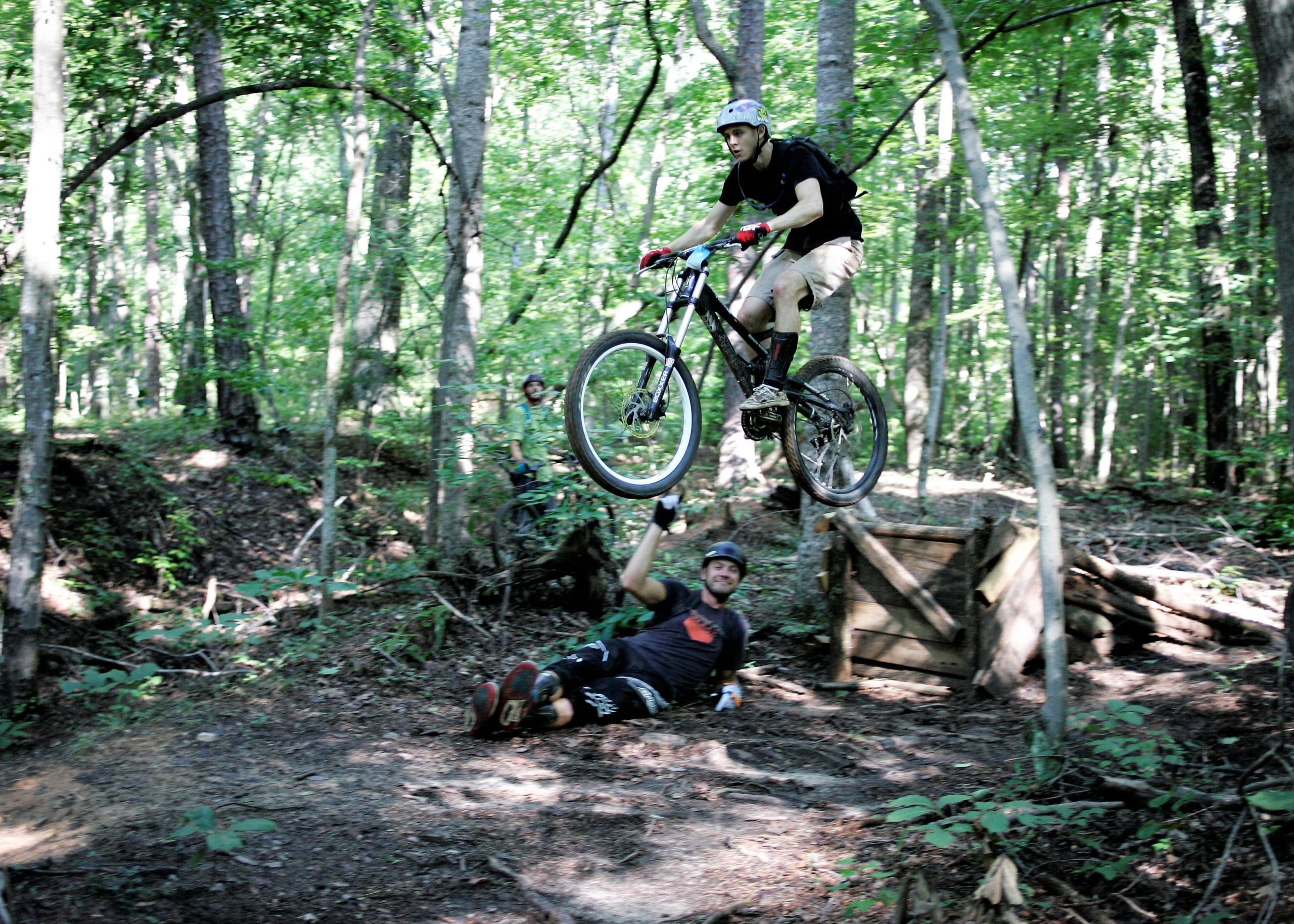 A mountain biker performing a jump over a wooden structure in a forested area, with a second rider sitting on the ground, smiling and giving a thumbs up in support. Surrounding trees create a vibrant green backdrop, showcasing a lively outdoor environment. Salem Lake mountain bike trail.