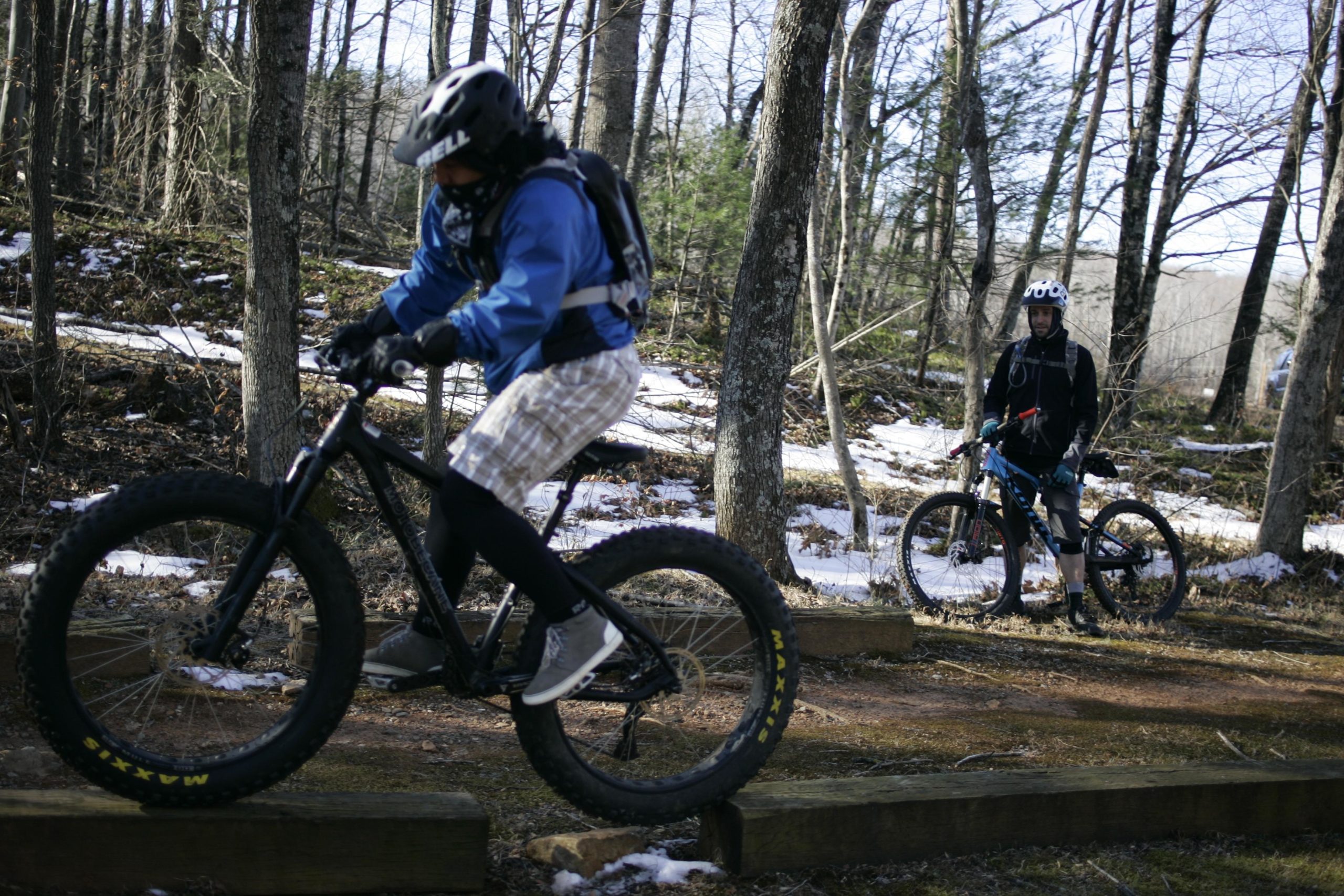 Two mountain bikers are riding through a wooded area with snow on the ground. One rider in a blue jacket and shorts is navigating over a wooden log, while the other rider, dressed in a dark outfit, stands beside their bike, observing the action. Trees and foliage surround the scene, indicating an outdoor trail. Woolwine Trails [Shiners Revenge] mountain bike trail.