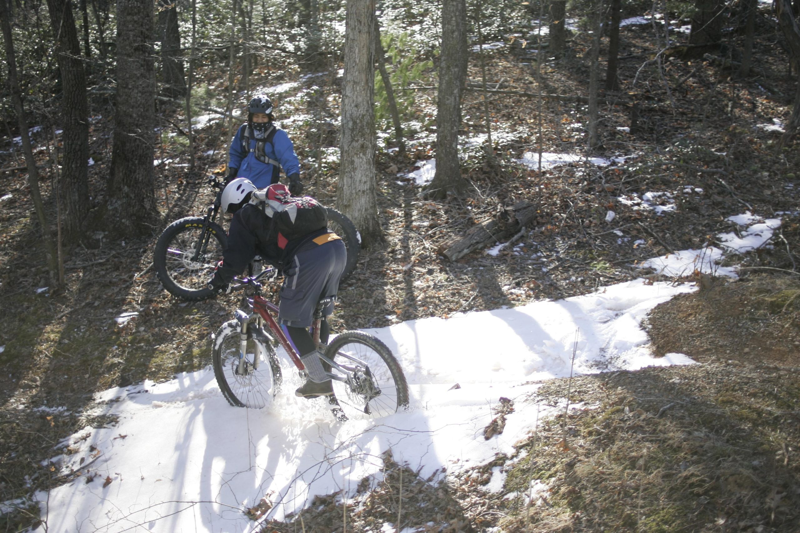 Two mountain bikers navigate a snowy trail in a wooded area. One rider is seen leaning forward on a bike, kicking up snow, while the other stands nearby with their bike, both dressed in winter gear. The scene features sunlight filtering through trees, illuminating patches of snow on the ground among fallen leaves and dirt. Woolwine Trails [Shiners Revenge] mountain bike trail.