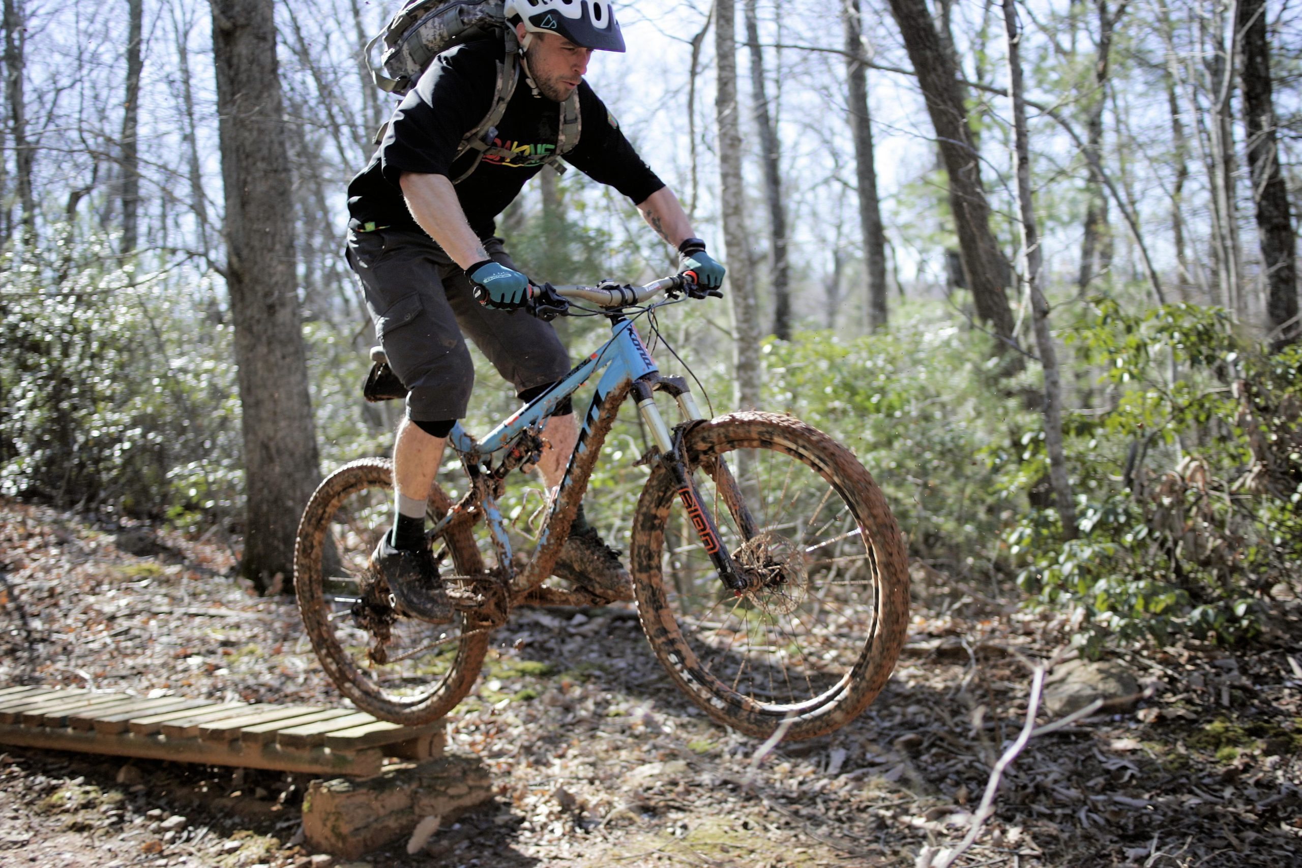 A mountain biker in mid-air jumps off a wooden bridge on a forest trail, showcasing an adventurous moment. The cyclist is dressed in a black shirt and shorts, wearing a helmet and gloves. His bike is splattered with mud, and the surrounding woods are filled with greenery and trees under a clear blue sky. Woolwine Trails [Shiners Revenge] mountain bike trail.