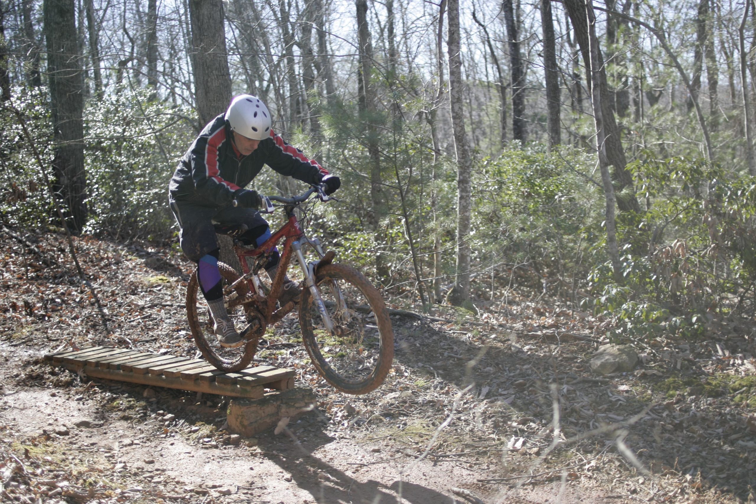 A mountain biker in a helmet and protective gear is mid-jump over a wooden bridge on a dirt trail surrounded by trees and vegetation. The biker's bicycle is partially airborne, with one wheel leaving the ground, while the terrain shows signs of mud and fallen leaves. Woolwine Trails [Shiners Revenge] mountain bike trail.