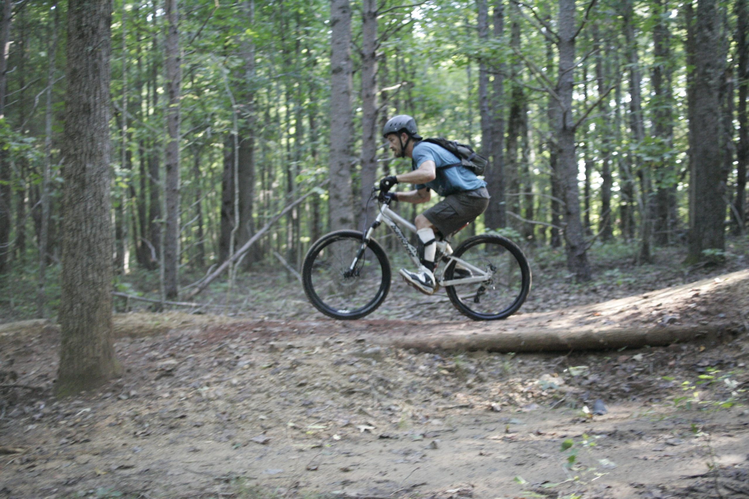 A person riding a mountain bike over a log on a dirt trail surrounded by dense green forest. The rider is wearing a helmet and riding gear, demonstrating an action-packed moment in outdoor biking. Angler's Ridge mountain bike trail.