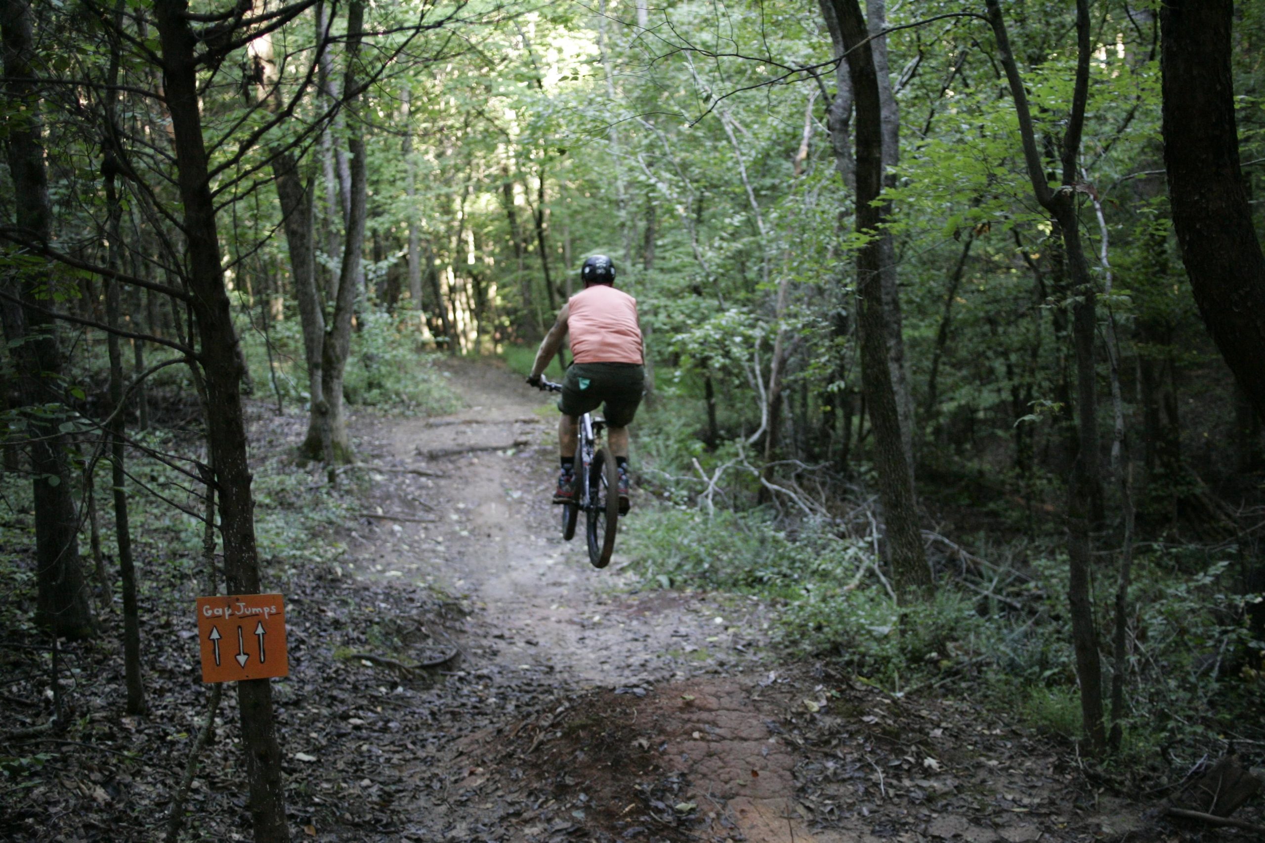 A mountain biker jumps off a dirt ramp on a forest trail, surrounded by green trees and foliage. An orange sign labeled "Gap Jumps" is visible on the left side of the path. Angler's Ridge mountain bike trail.