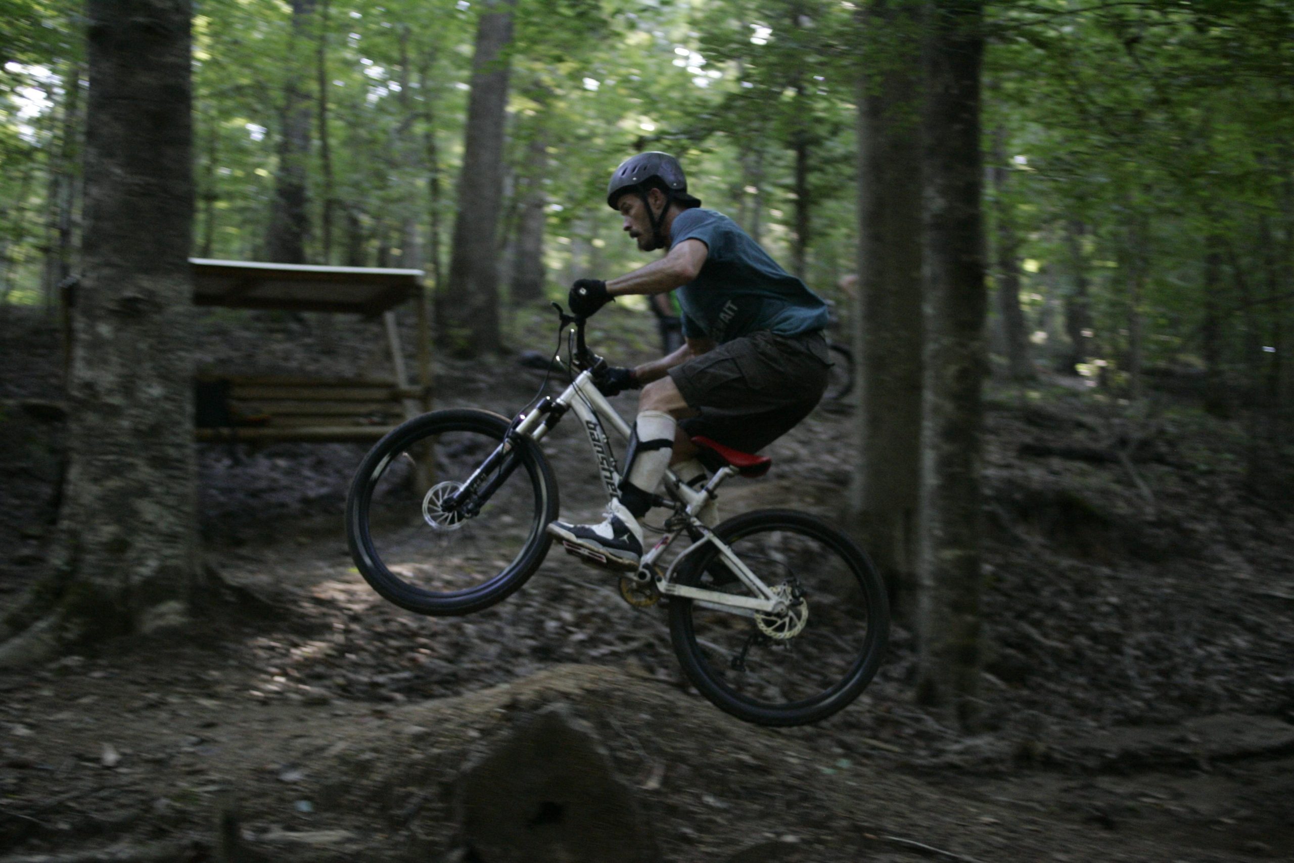 A mountain biker performing a jump on a trail in a wooded area, surrounded by trees and foliage. The biker is wearing a helmet and riding a white bicycle, with both wheels off the ground as he navigates over a dirt mound. Angler's Ridge mountain bike trail.