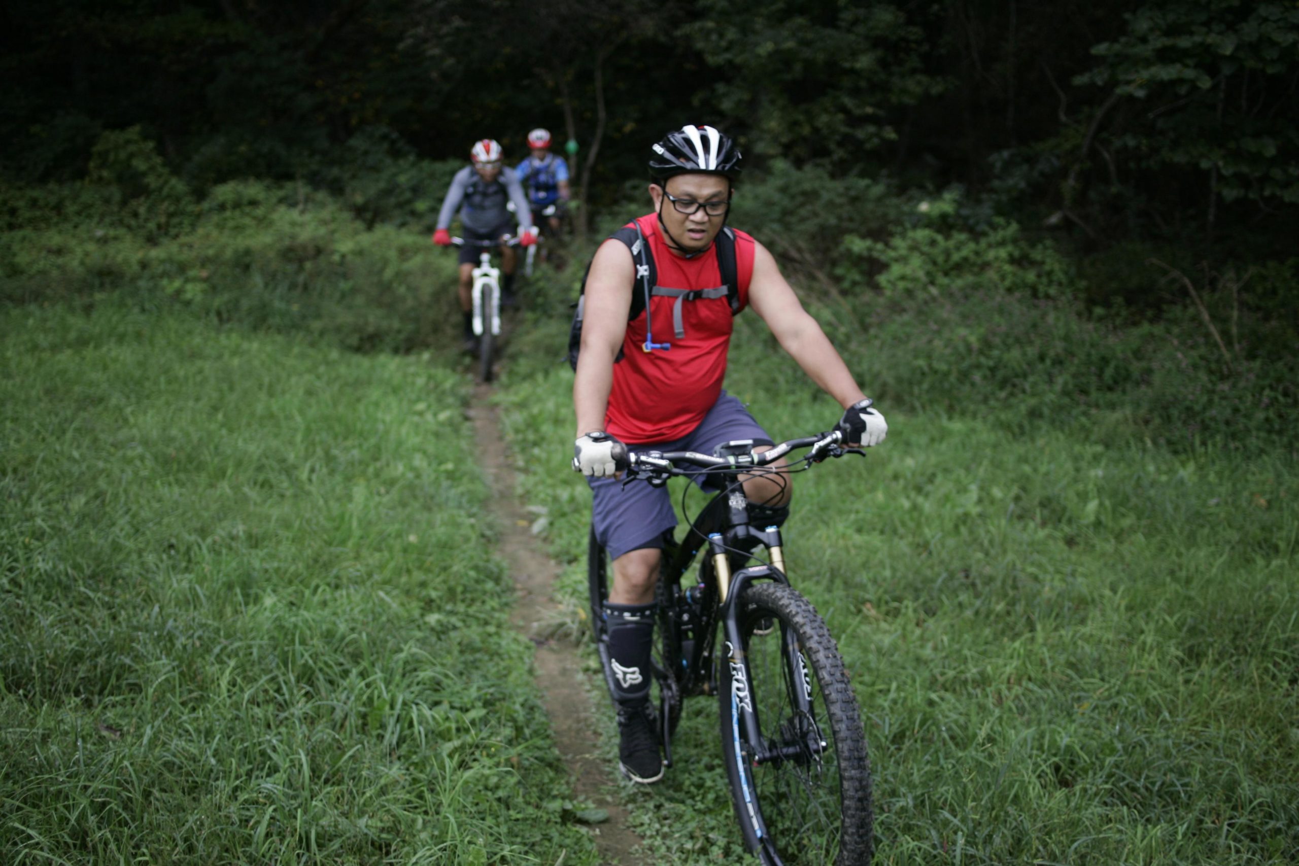 A group of three mountain bikers riding on a narrow dirt path through lush green grass and foliage. The forefront rider wears a red tank top and a helmet, focused on the trail ahead, while two other riders can be seen in the background. Massanutten Western Slope mountain bike trail.