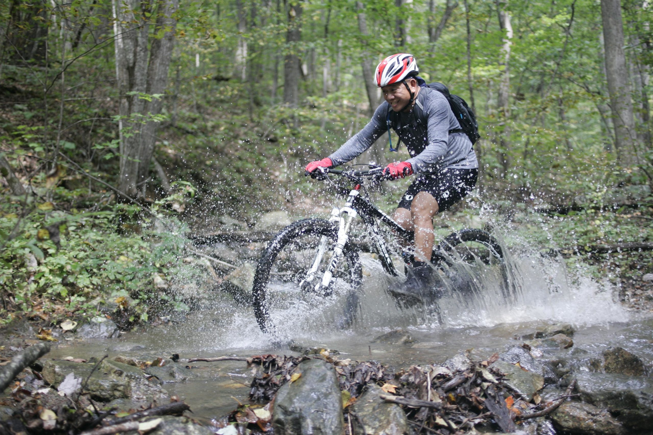 A person riding a mountain bike through a stream in a forested area, splashing water as they navigate over rocks. The cyclist is wearing a helmet, gloves, and a long-sleeved shirt, with greenery and trees surrounding the scene. Massanutten Western Slope mountain bike trail.