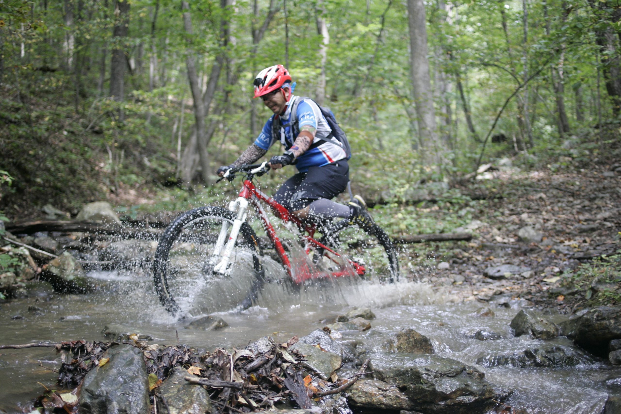 A mountain biker navigating through a rocky creek in a forested area, splashing water as they ride. The biker is wearing a helmet and a blue jersey, with trees and greenery surrounding the scene. Massanutten Western Slope mountain bike trail.