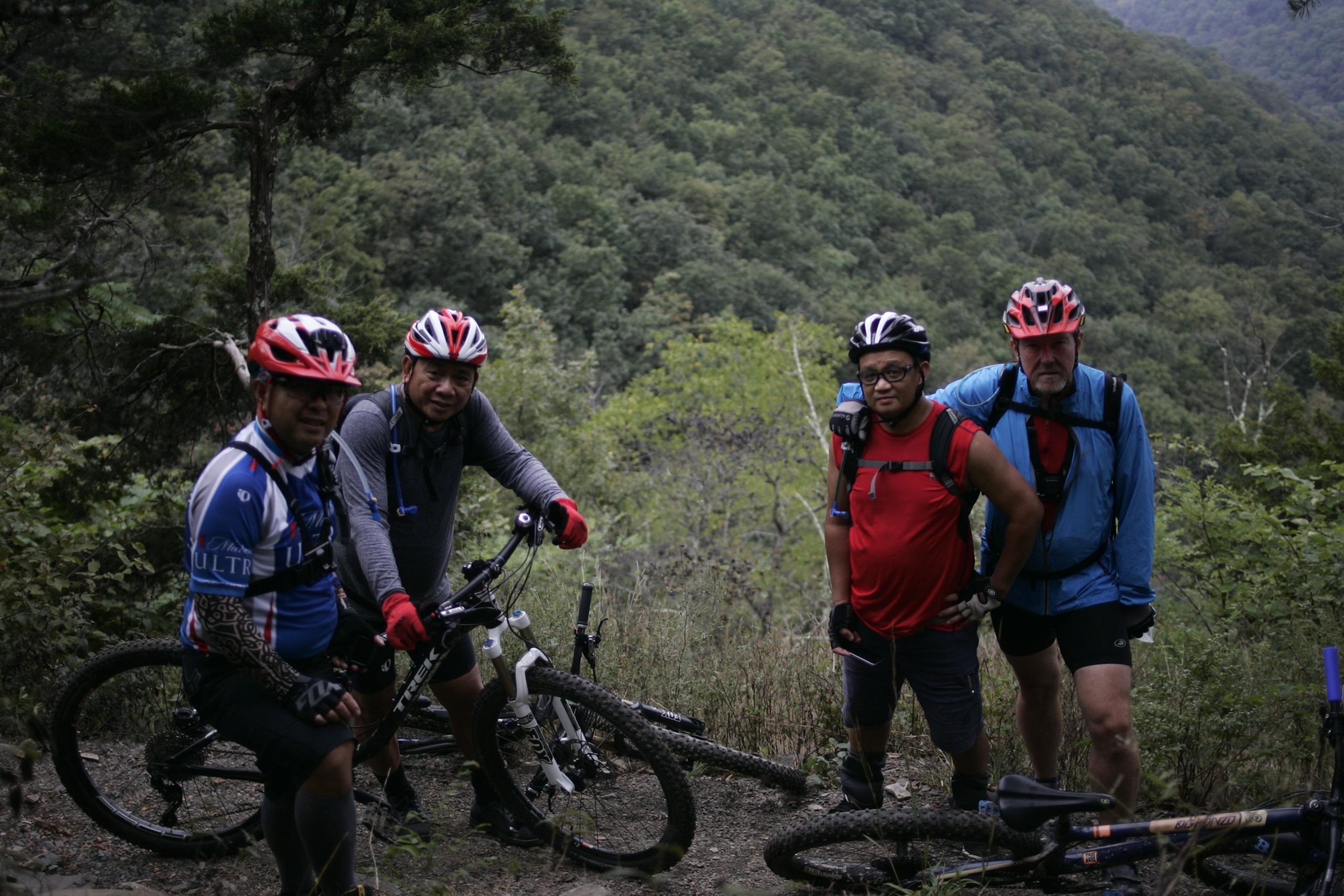 Four mountain bikers pause on a trail in a forested area, surrounded by lush greenery and rolling hills. Two riders are positioned closer to the camera, wearing helmets and cycling gear, while the other two stand slightly further back. One bike is propped upright against a rock, and the group appears relaxed and engaged in conversation, enjoying their outdoor adventure. Massanutten Western Slope mountain bike trail.