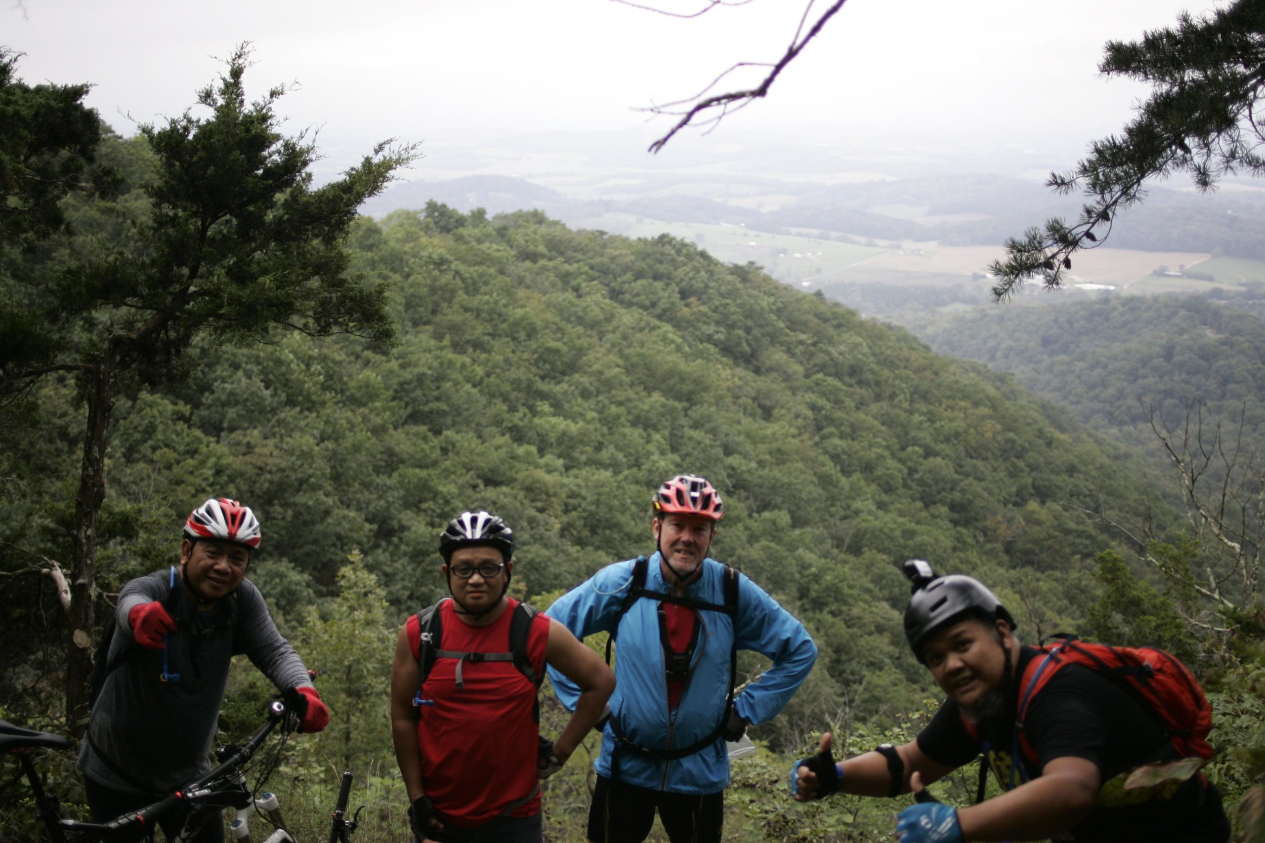 Four mountain bikers pose for a photo on a hillside overlooking a lush, green valley. They wear helmets and biking gear, with two of them giving thumbs-up signs. The background features rolling hills and a cloudy sky, suggesting an adventurous outdoor setting. Massanutten Western Slope mountain bike trail.