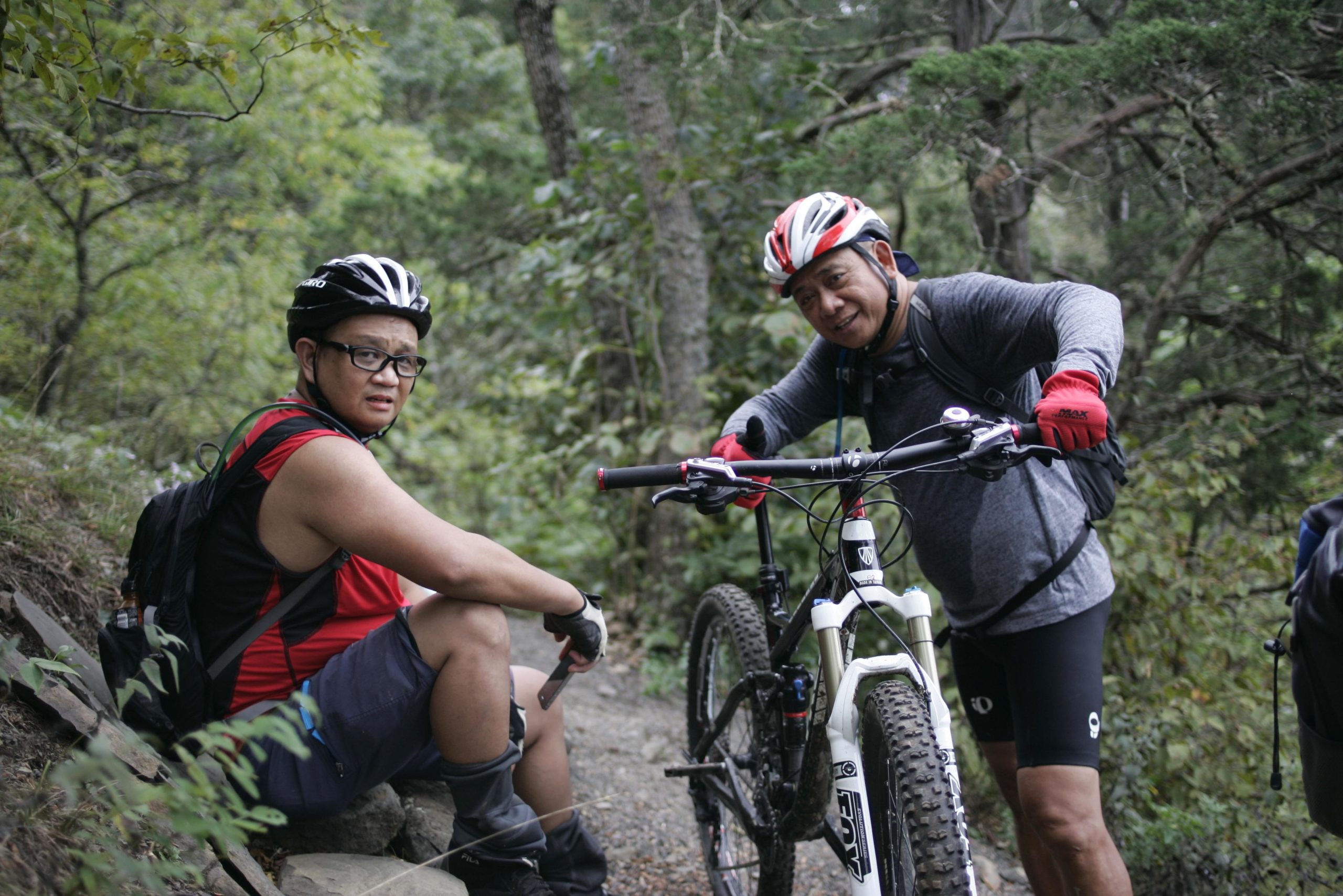 Two mountain bikers are positioned on a trail surrounded by lush greenery. One rider, wearing a red sleeveless shirt and glasses, is seated on a rock, looking towards the camera with a thoughtful expression. The other rider, dressed in a gray long-sleeve shirt and red gloves, is standing nearby with a mountain bike, smiling while holding onto the handlebars. The setting suggests a break in an outdoor biking adventure. Massanutten Western Slope mountain bike trail.