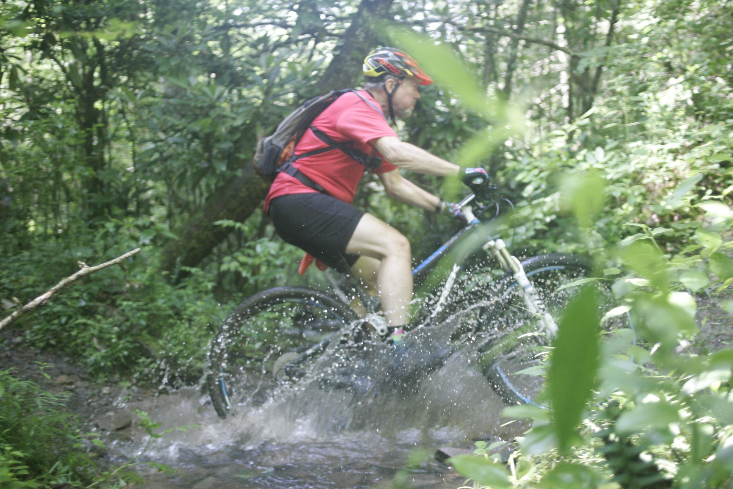 A cyclist wearing a red shirt and helmet rides through a muddy trail in a lush green forest, splashing water as they navigate over a puddle. Tsali Recreation Area mountain bike trail.