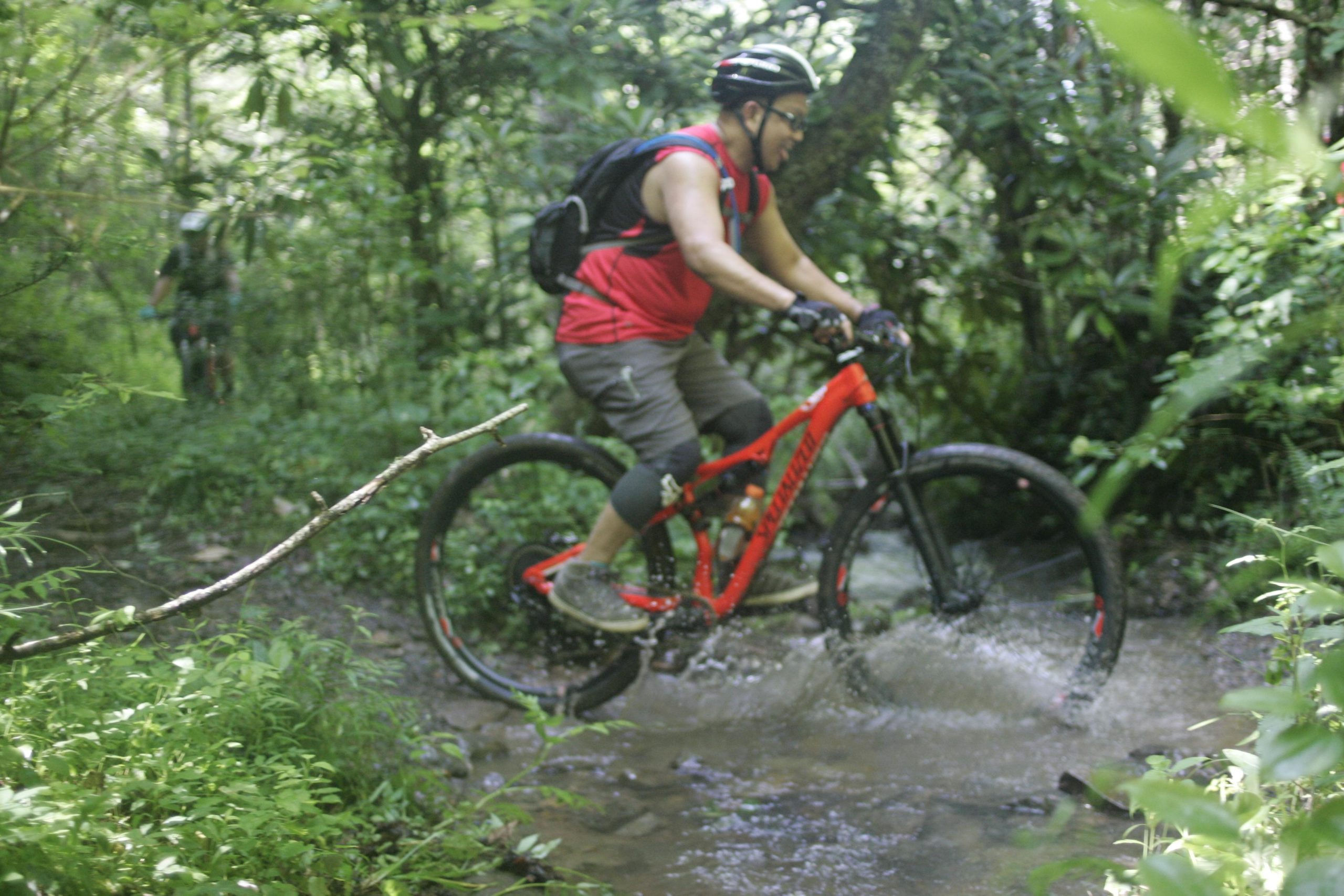 A person riding a mountain bike through a shallow stream on a lush, green trail surrounded by trees. The cyclist, wearing a red shirt, helmet, and protective gear, smiles as they navigate the water, with splashes occurring around the bike. A second cyclist is faintly visible in the background. Tsali Left Loop mountain bike trail.